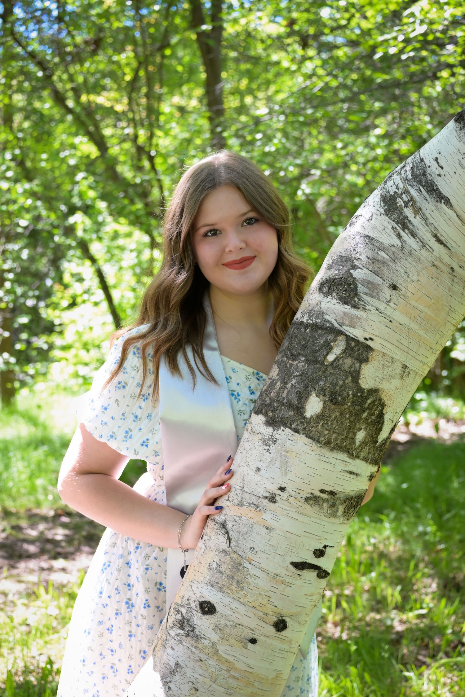 A person in a floral dress poses next to a white birch tree trunk in a sunlit forest setting.