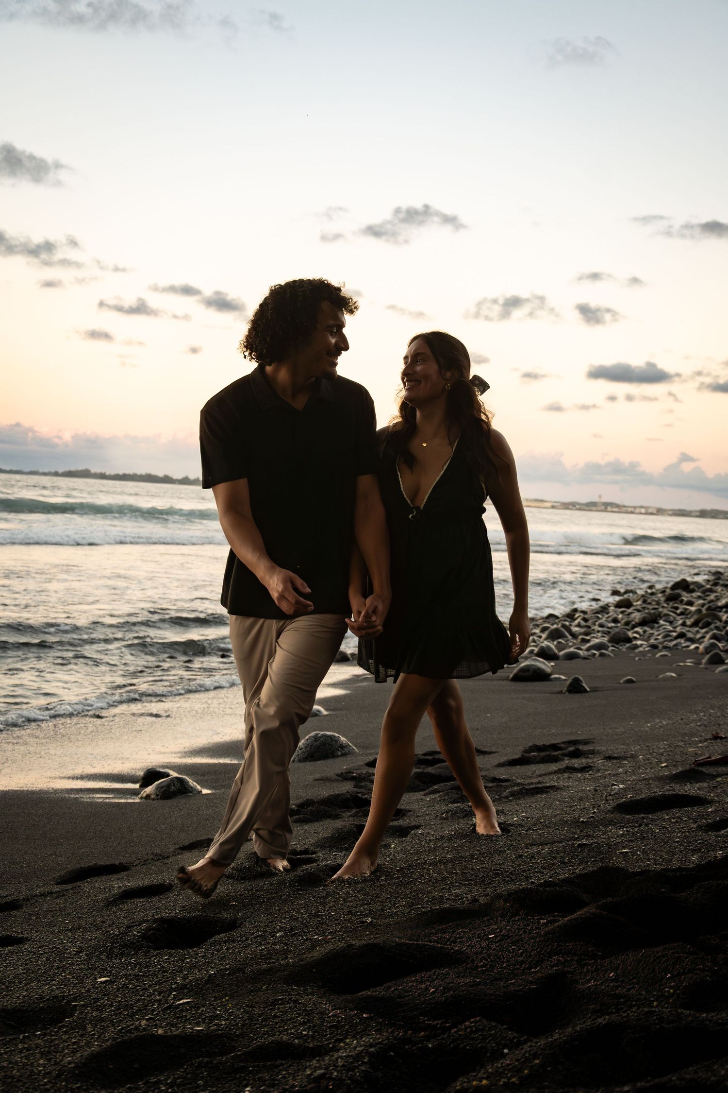Two figures walk together on a black sand beach at sunset with waves crashing in the background.