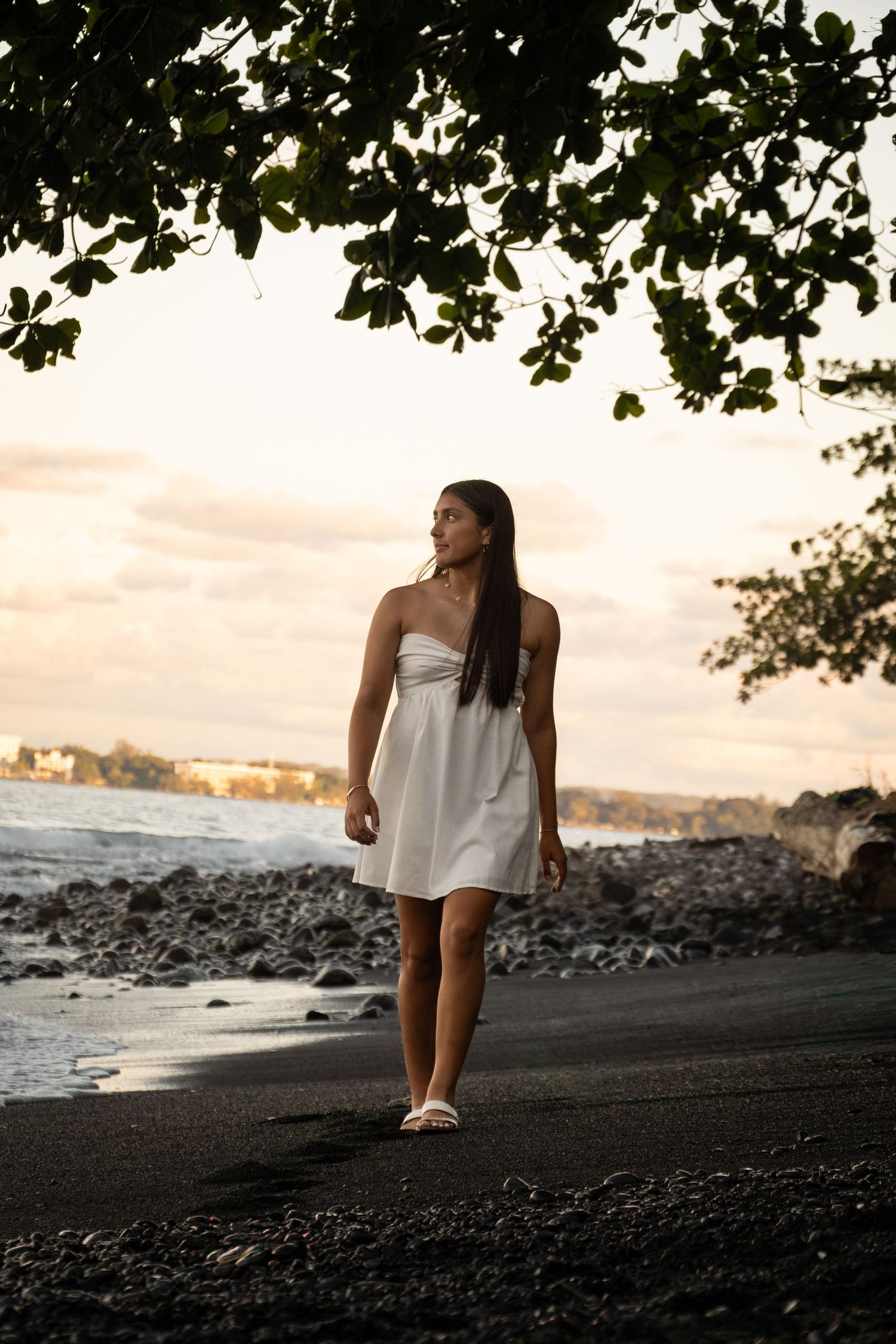 Person in white dress walks along black sand beach at sunset with trees overhead.