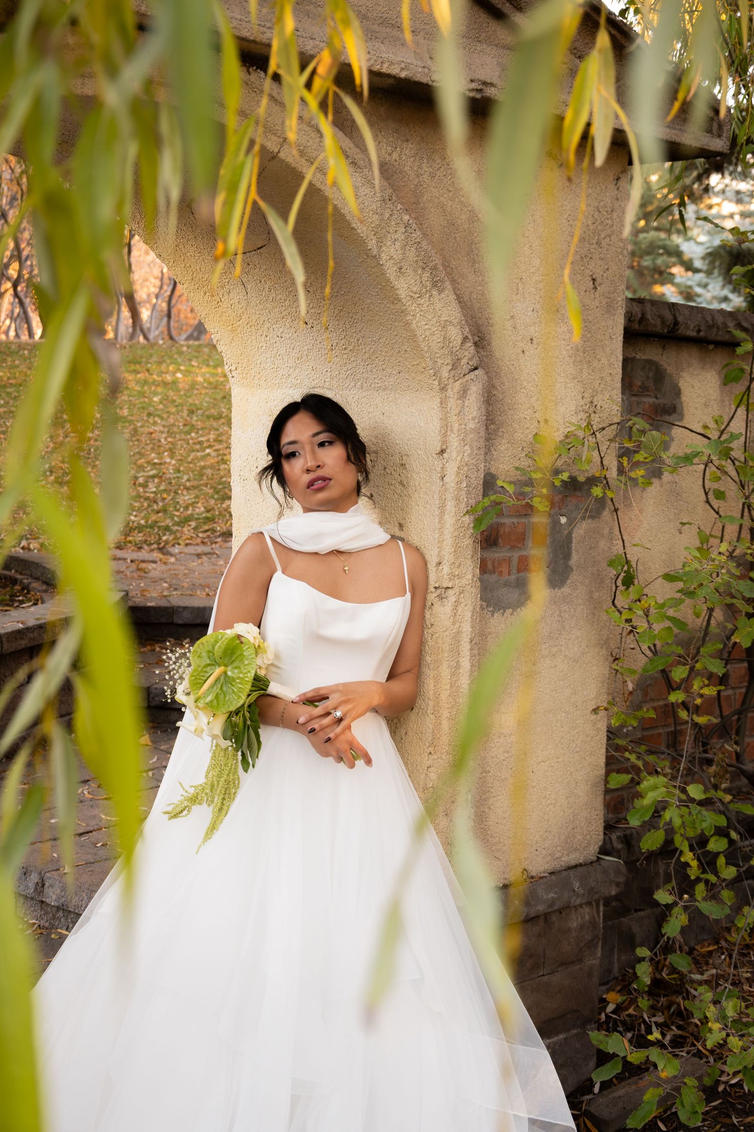 Beautiful bride in white ball gown with scarf neckline holding bouquet under stone arch surrounded by greenery.