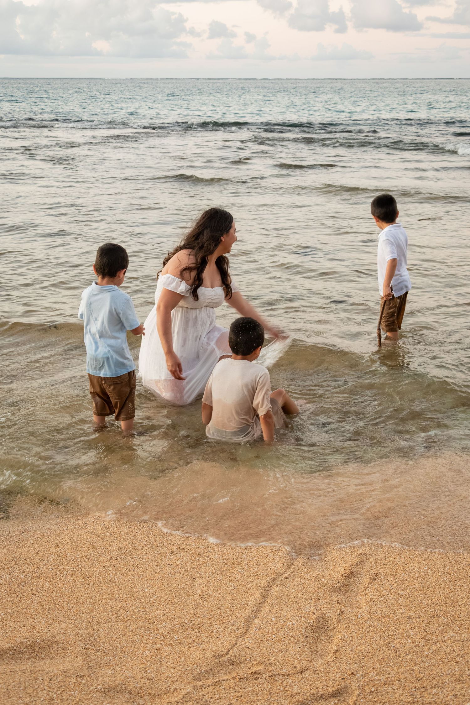 A mother in a white dress plays with her three young children in the shallow ocean waves on a sandy beach.