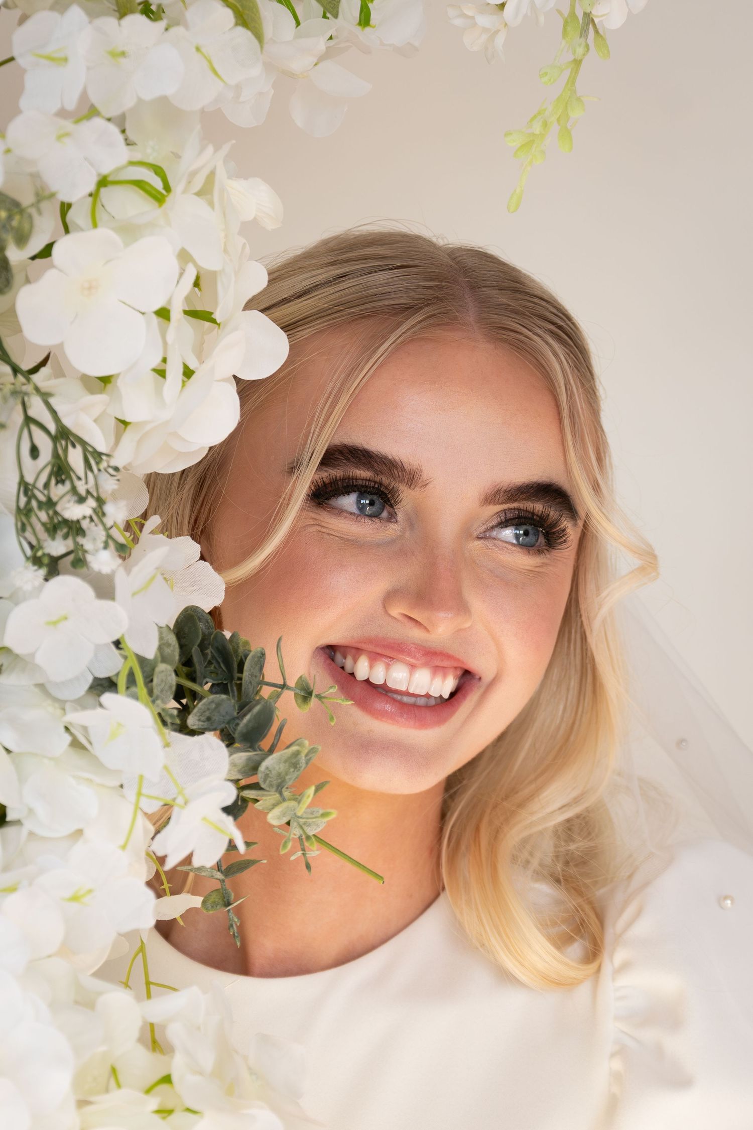 Smiling blonde bride with blue eyes surrounded by white flowers and greenery, wearing a veil and white dress.