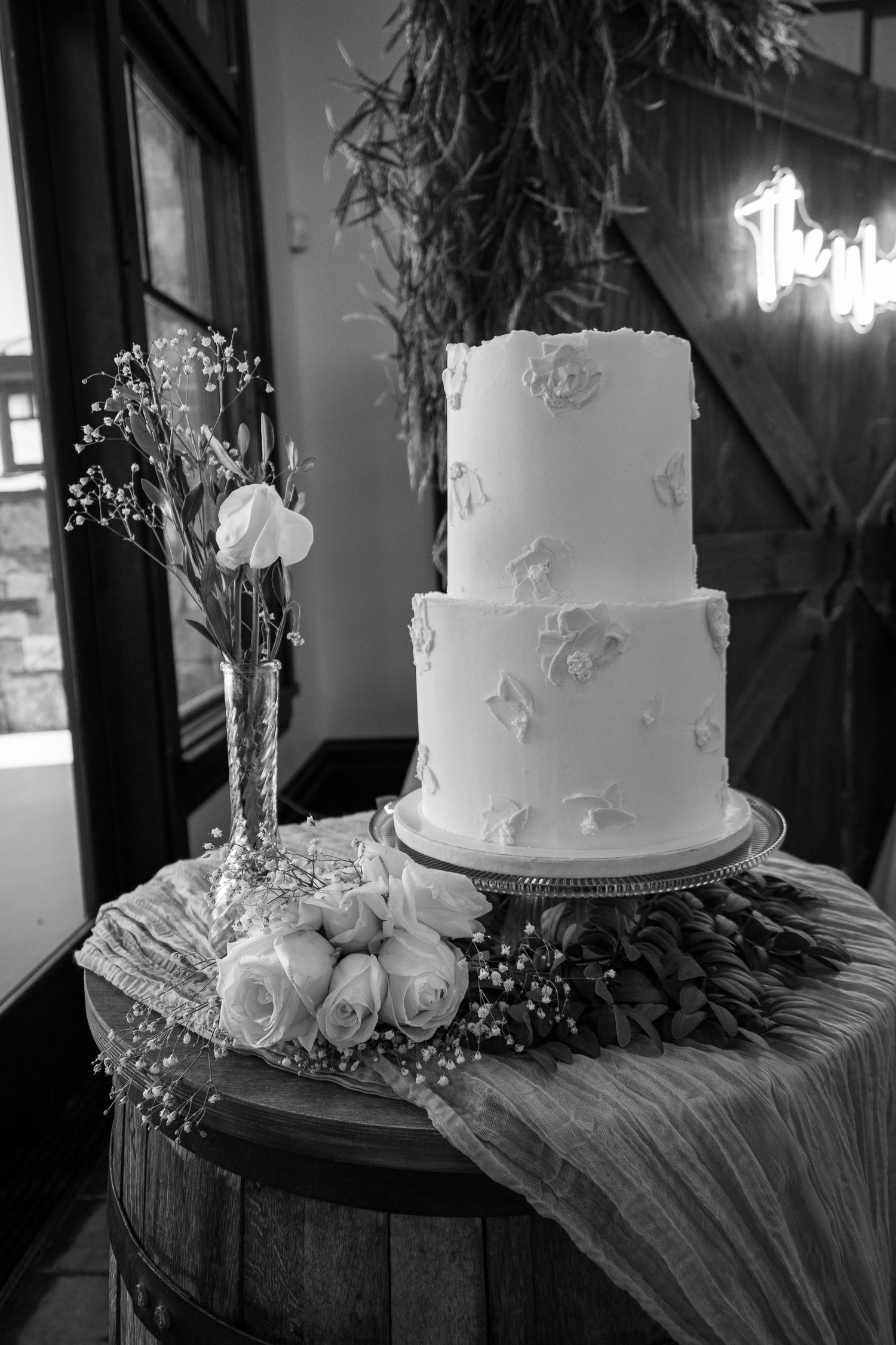Black and white photo of elegant two-tier white wedding cake with floral details on barrel table with roses and baby's breath.