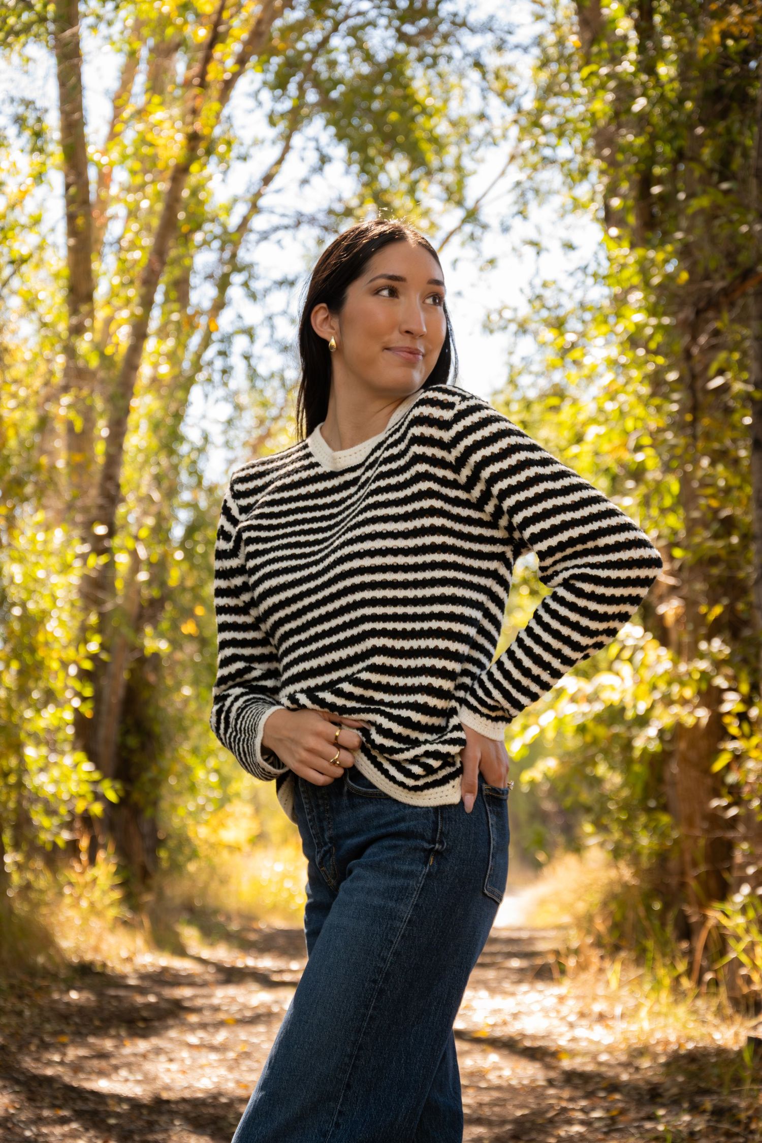 Person in striped sweater and jeans poses against autumn trees with golden sunlight filtering through the branches.