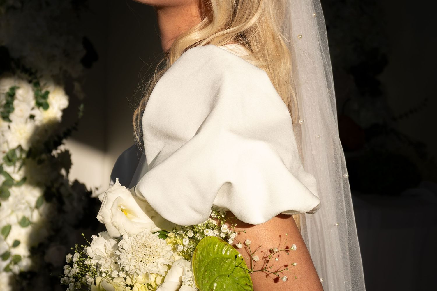 Bride in white peplum wedding dress holding bouquet with veil, floral tattoo visible on back.