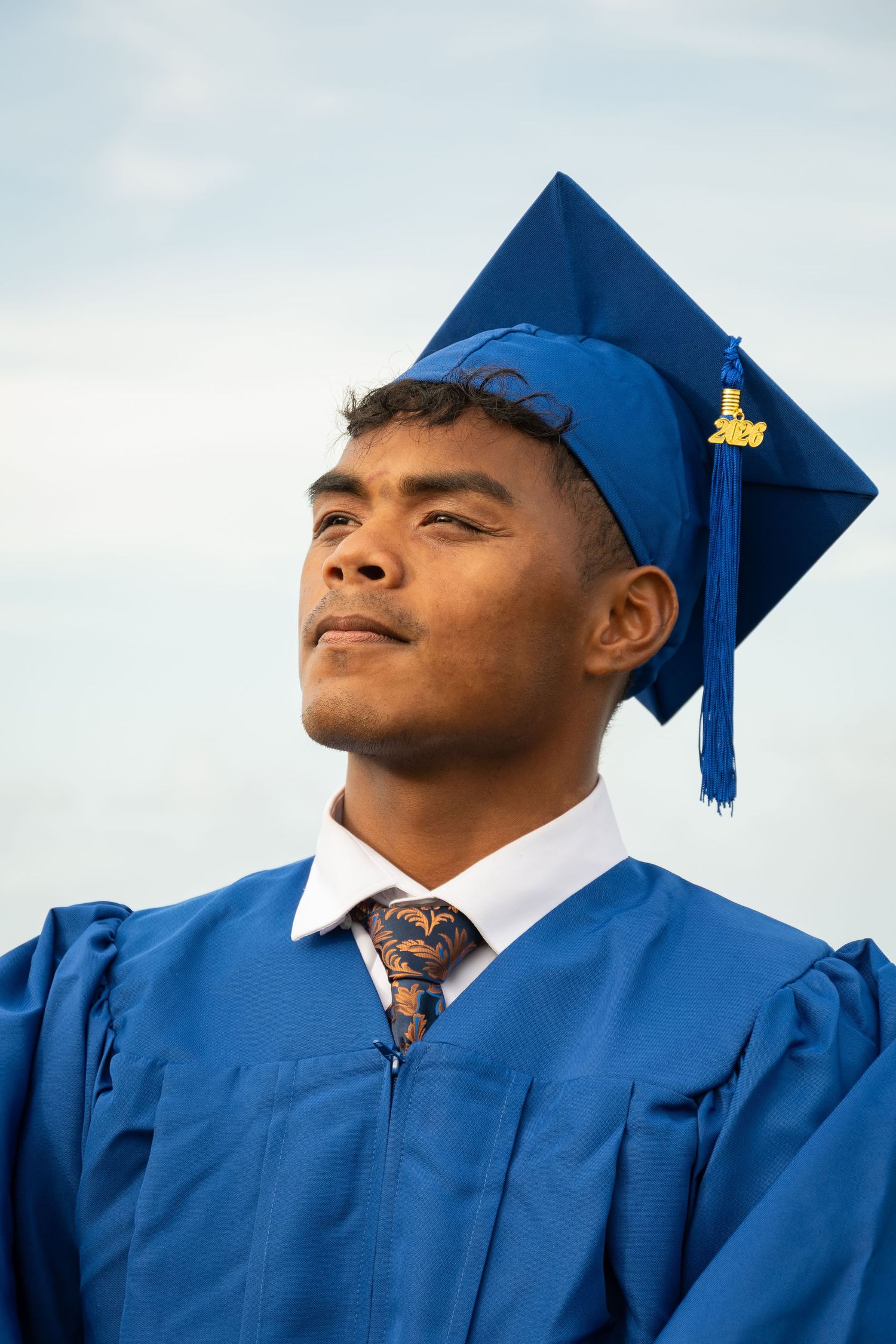 Young man in blue graduation cap and gown with decorative tie looks upward confidently against a cloudy sky.