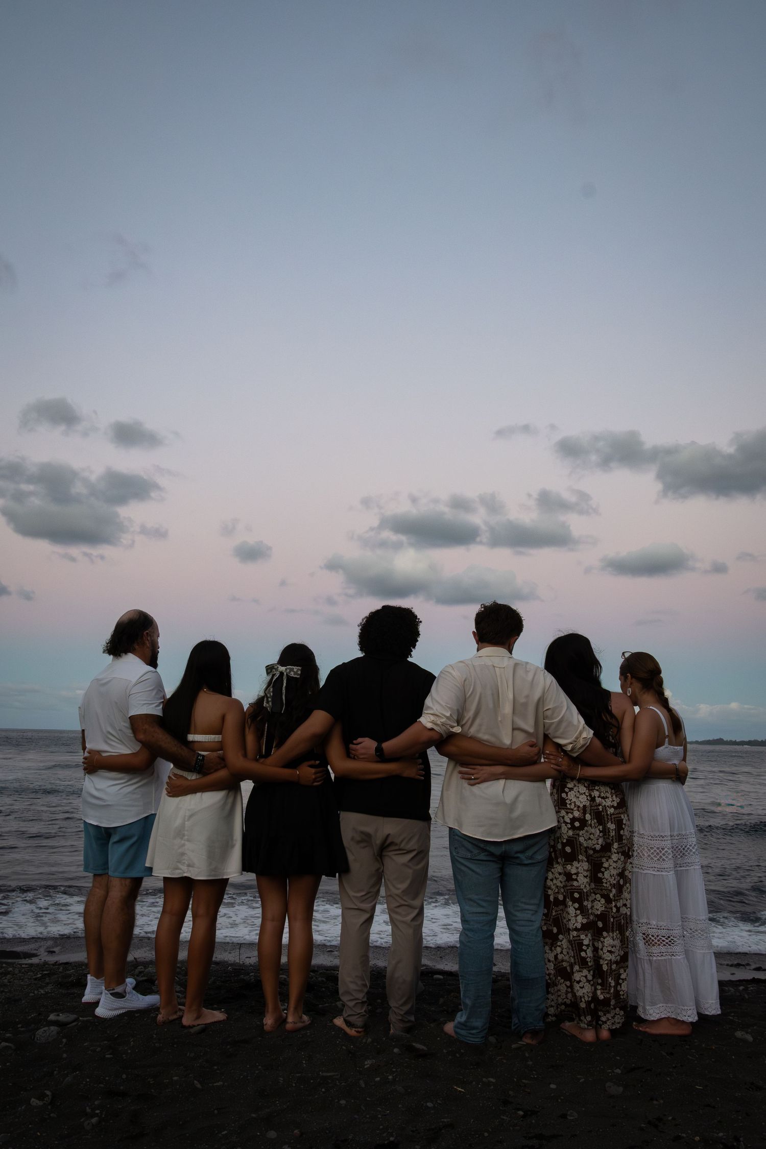A group of people stand together in silhouette against a sunset sky while looking out at the ocean.