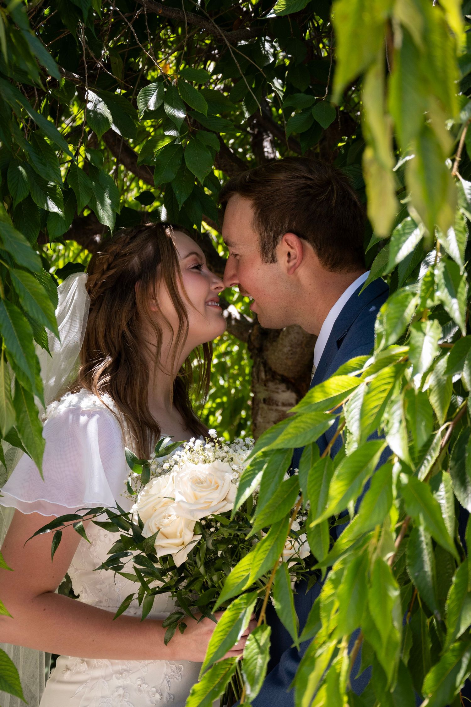 Romantic wedding couple sharing intimate moments under cascading willow branches with white floral bouquet.