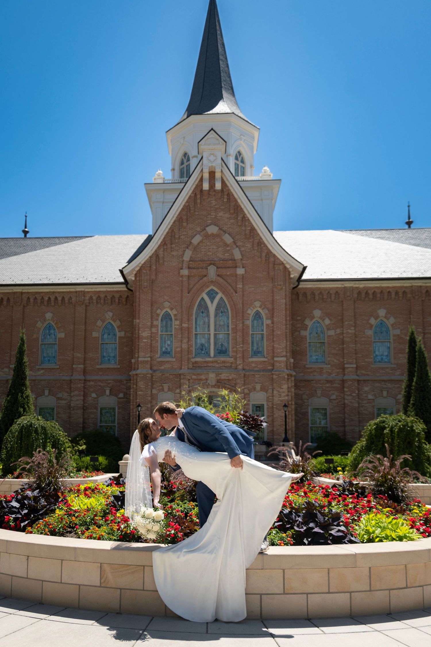A couple shares a romantic dip kiss in front of a beautiful brick church with white steeple and colorful flower garden.