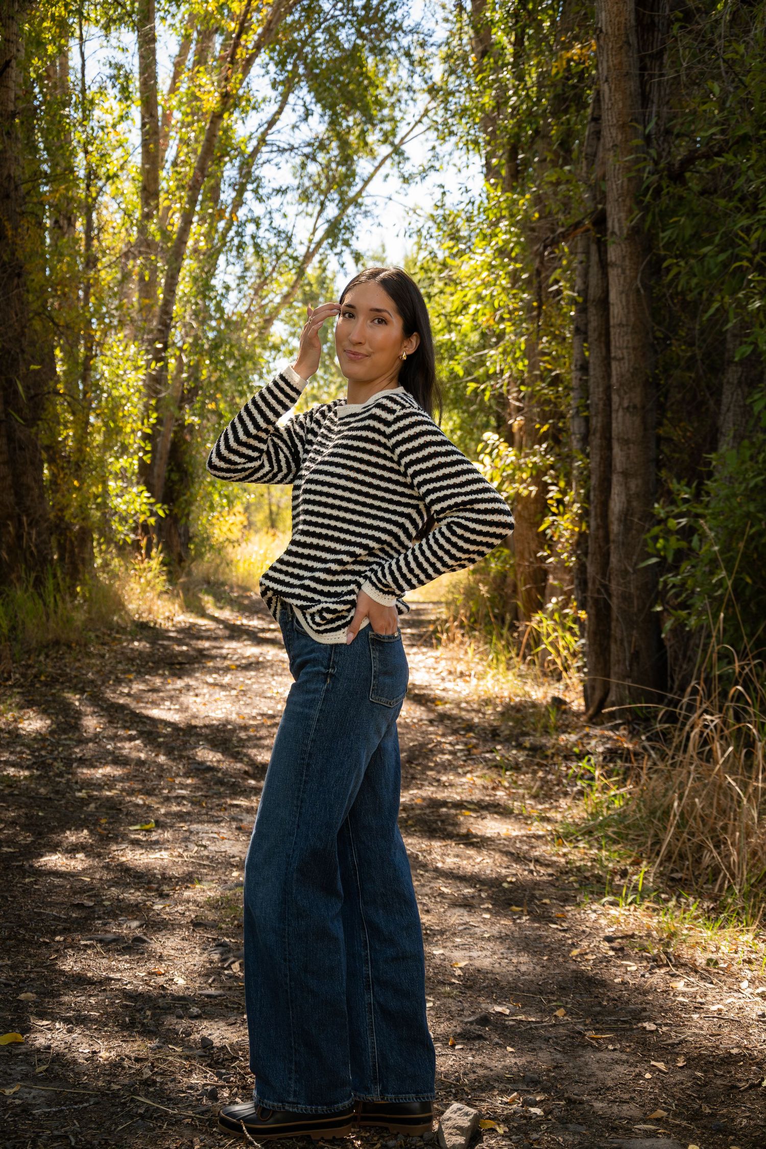 A person in a striped sweater and wide-leg jeans poses on a sunlit forest trail surrounded by autumn trees.