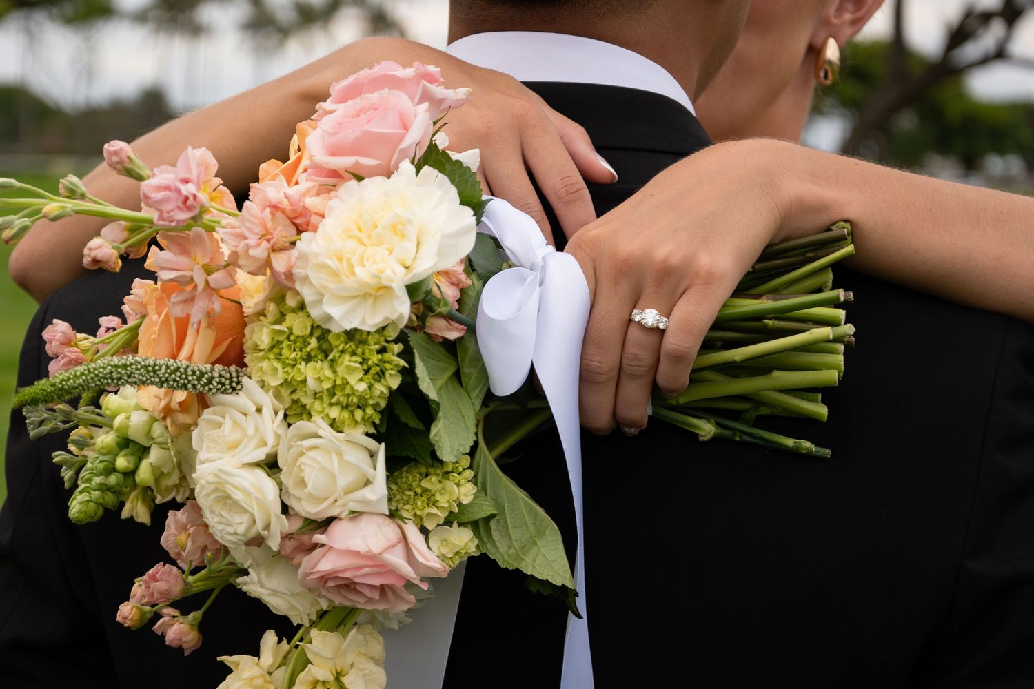 Bride holding a lush pink and white wedding bouquet while embracing groom in black suit, wearing diamond engagement ring.