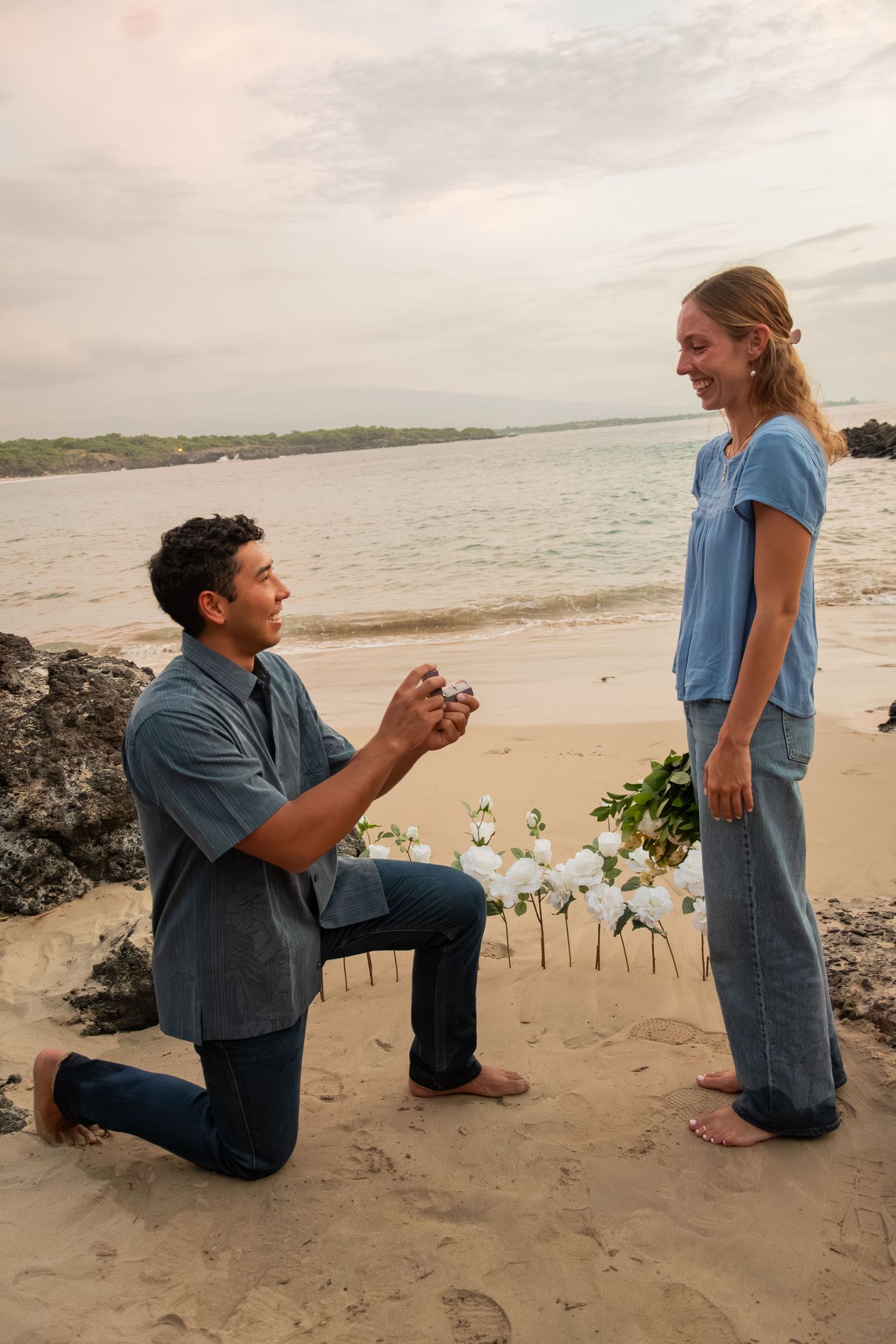 Man proposes on one knee to woman on sandy beach at sunset, holding ring with white flowers nearby.