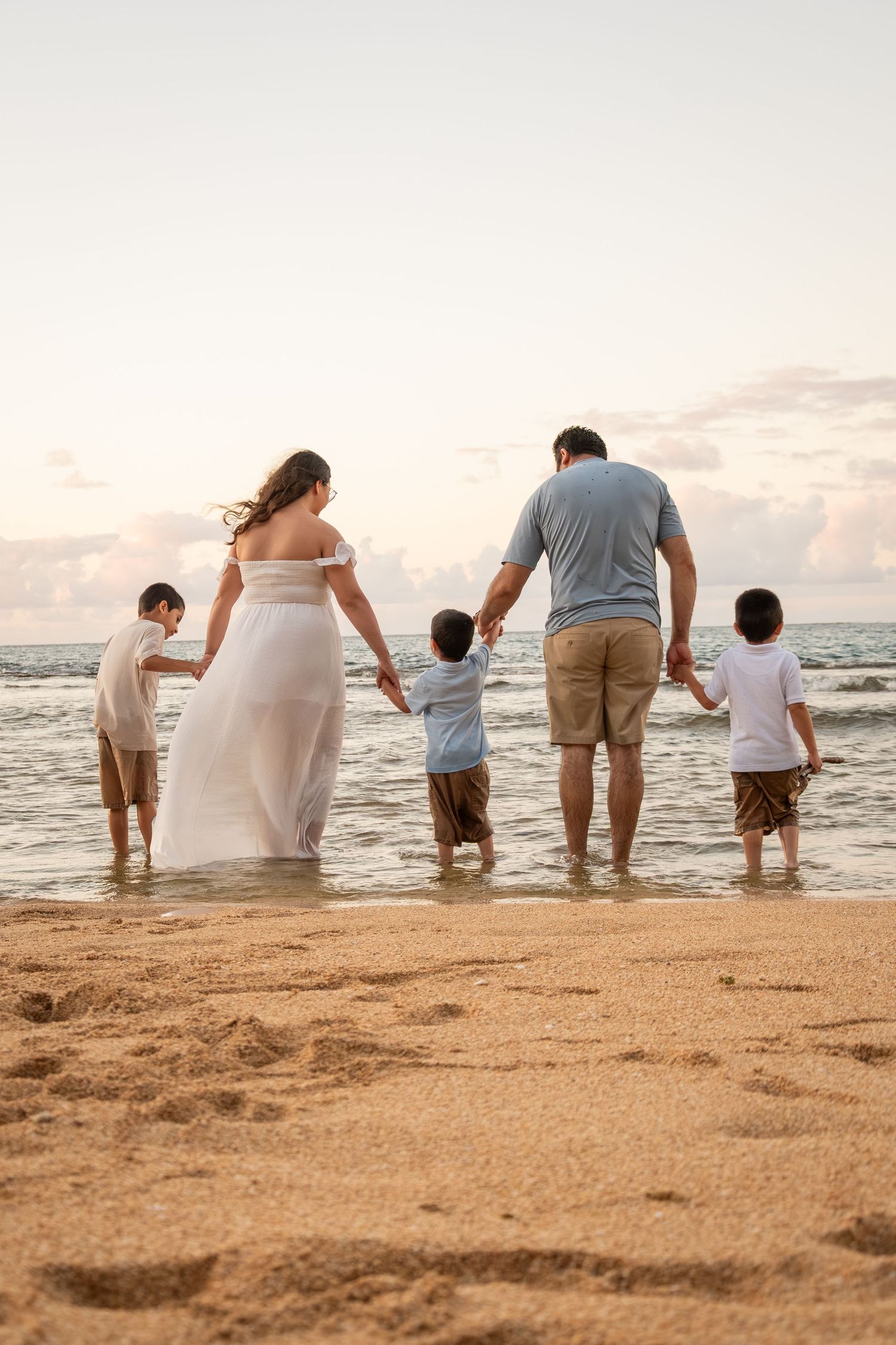 Family of five holding hands at the beach during sunset, parents and three young boys wading in ocean waves.