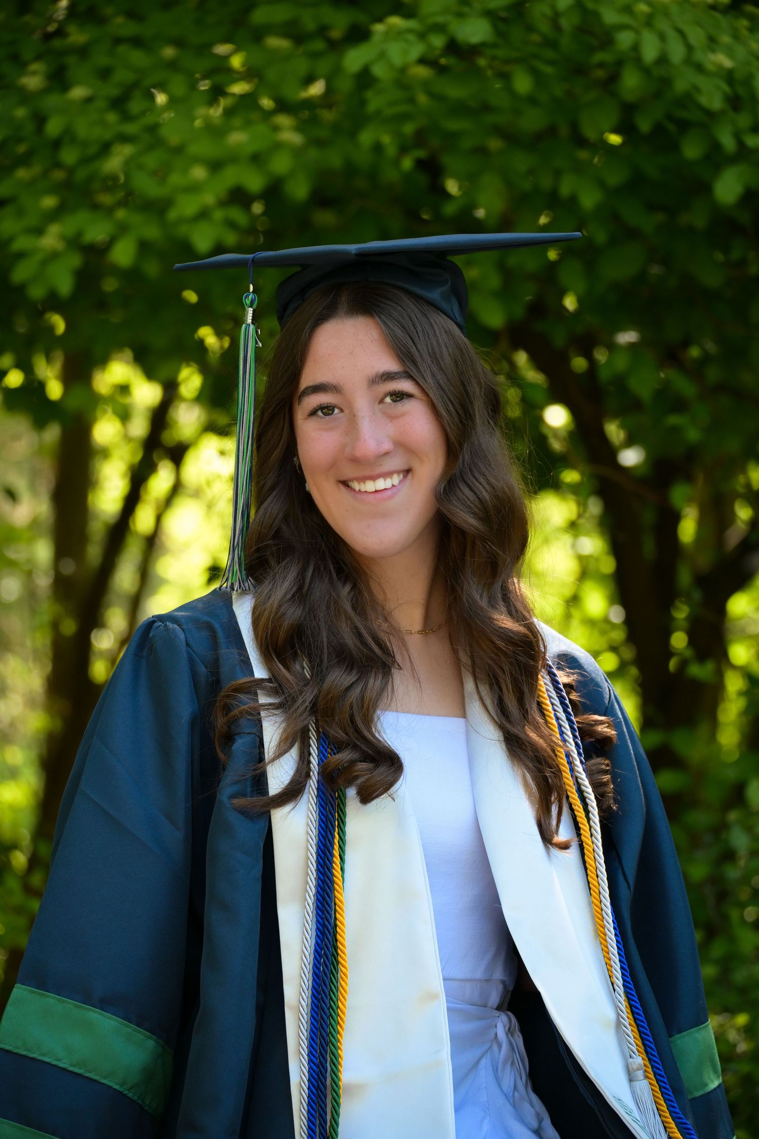 Graduate in green cap and gown poses for portraits in front of trees on sunny graduation day.
