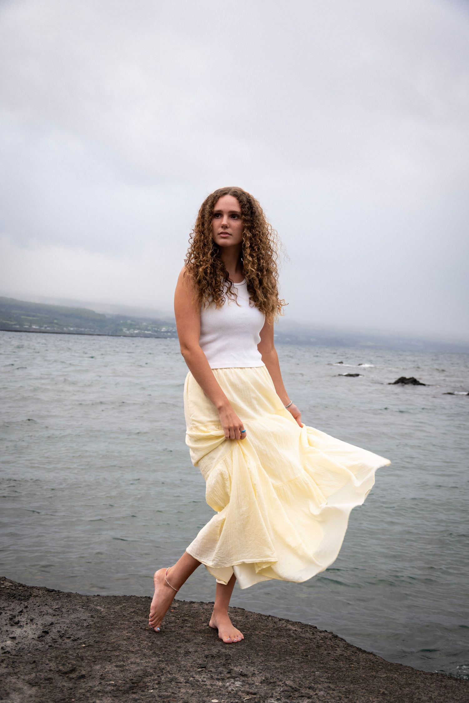 A flowing yellow dress moves gracefully against a misty coastal backdrop with rocks and ocean waves.