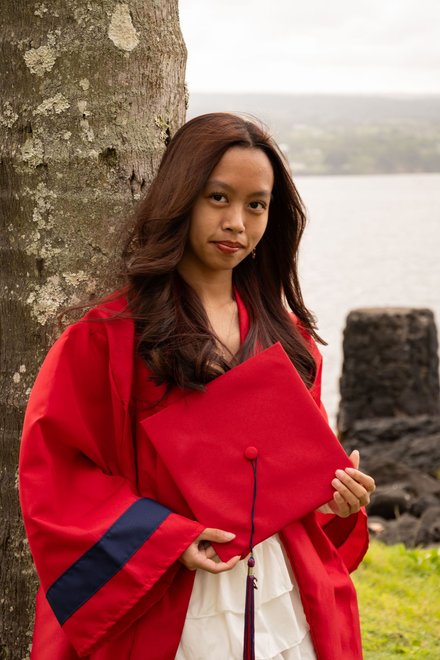 Young woman in red graduation gown holding cap, leaning against tree with water in background.