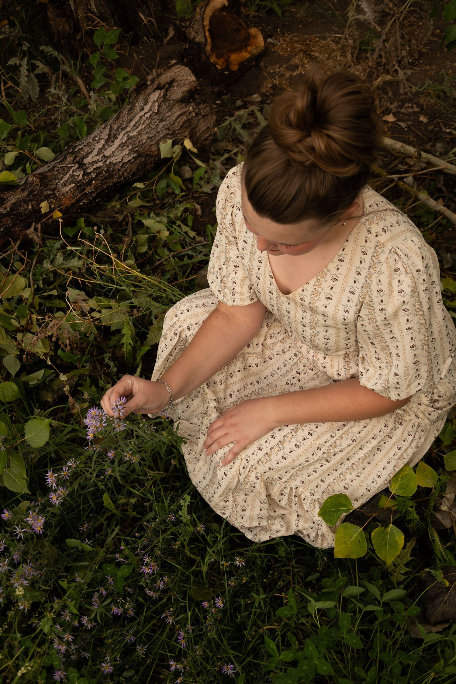 Woman in vintage floral dress sitting outdoors picking wildflowers near a fallen log.