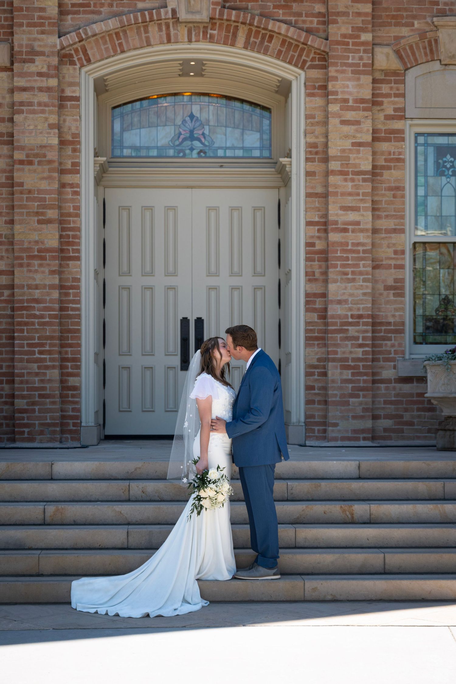A couple in wedding attire share a tender moment on brick steps in front of a vintage cream-colored door with stained glass.