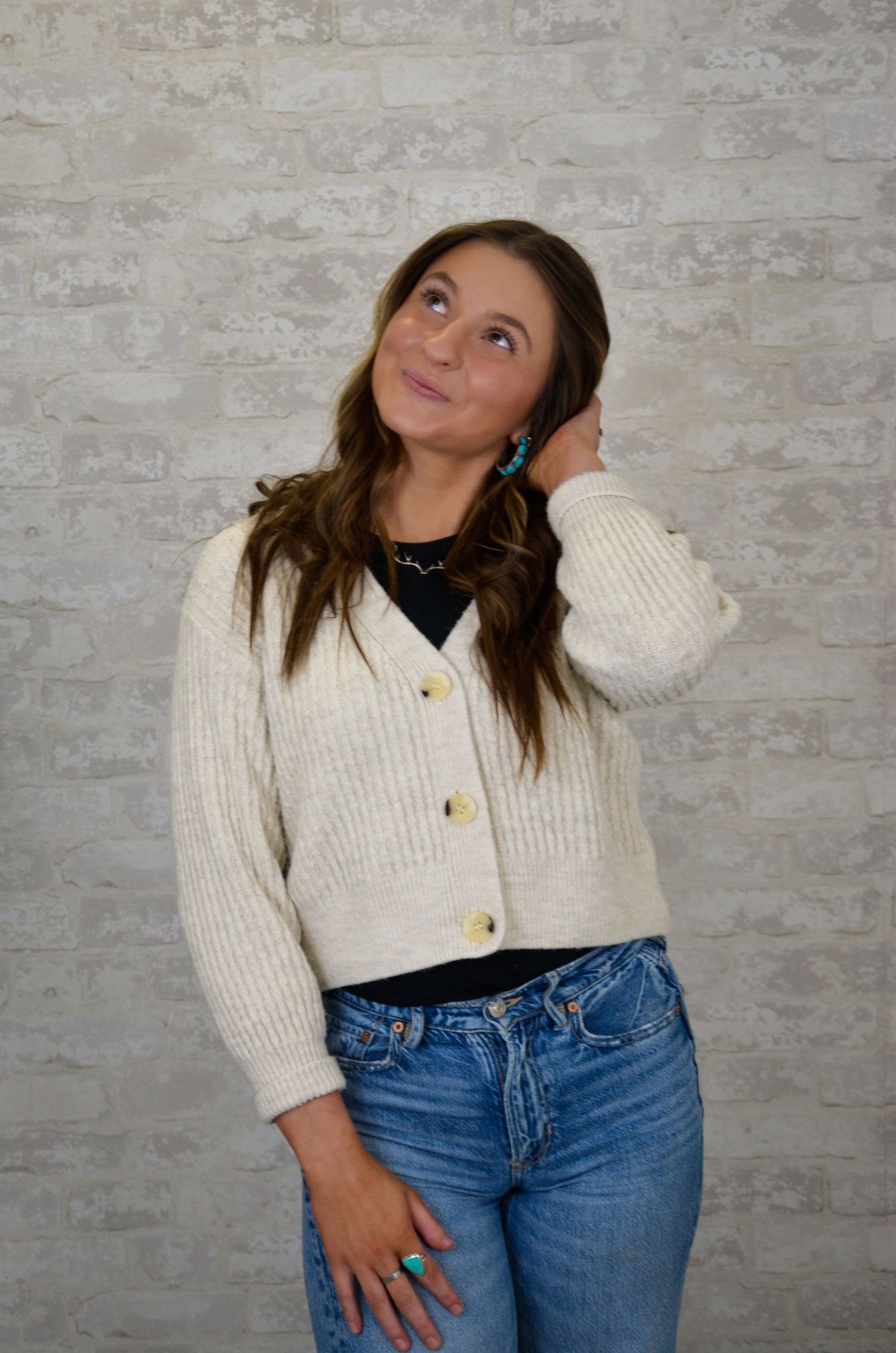 Person in white cardigan and jeans poses thoughtfully against brick wall.