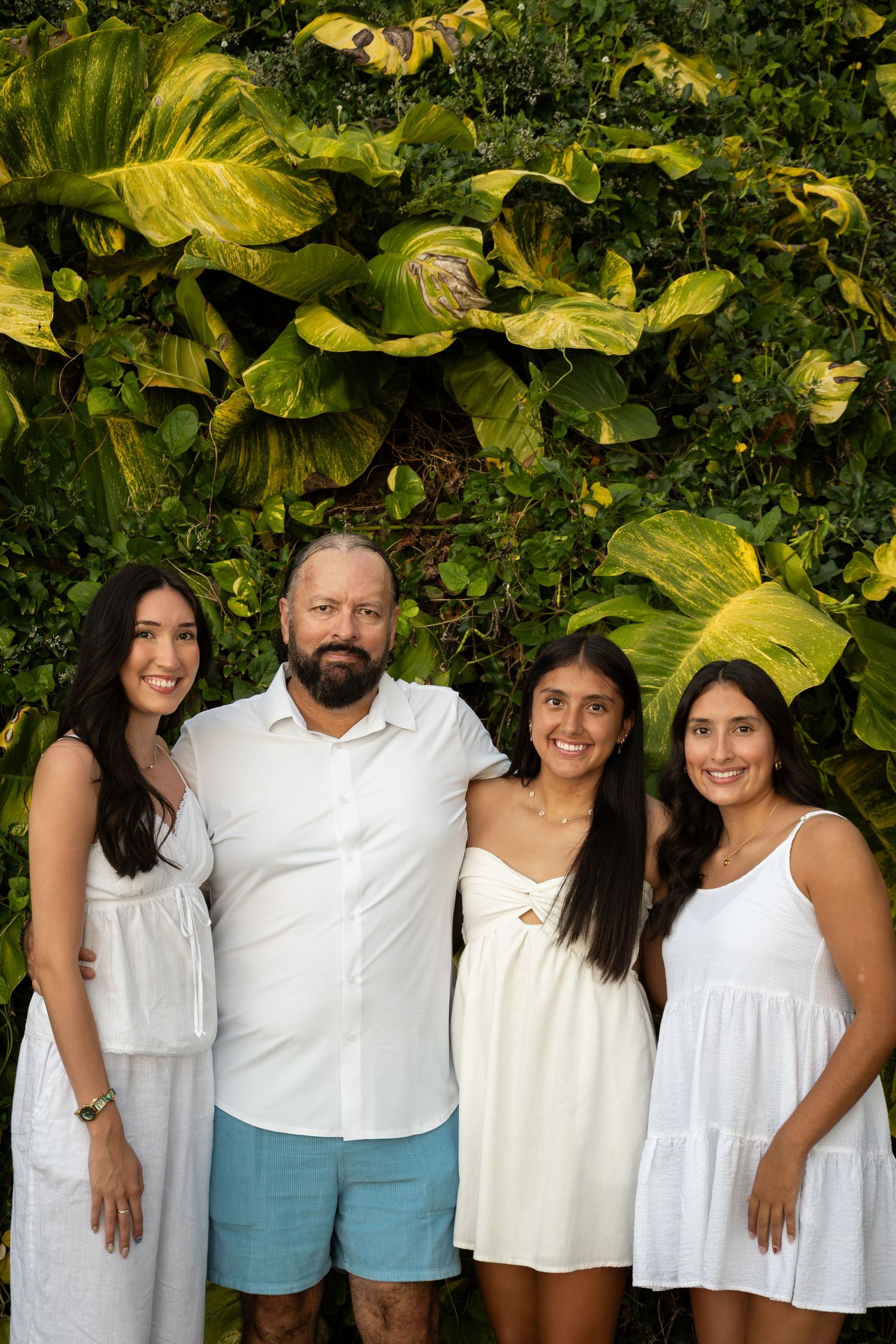 Family group wearing coordinated white outfits poses together against a backdrop of large tropical foliage.