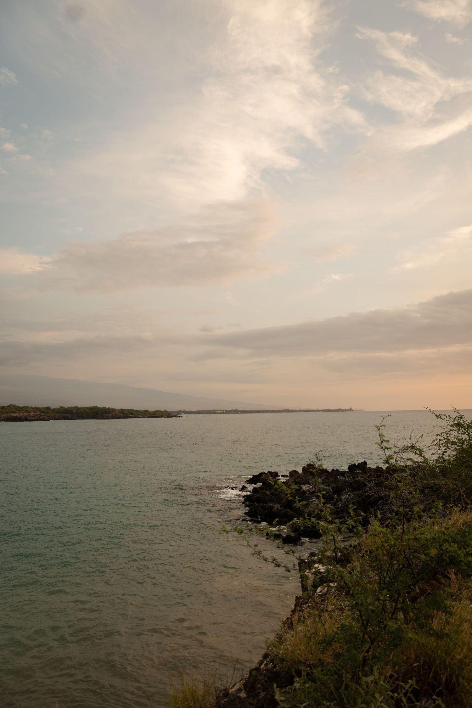 Serene coastal landscape with rocky shoreline, lush greenery, calm turquoise waters, and dramatic cloudy sky at dusk.