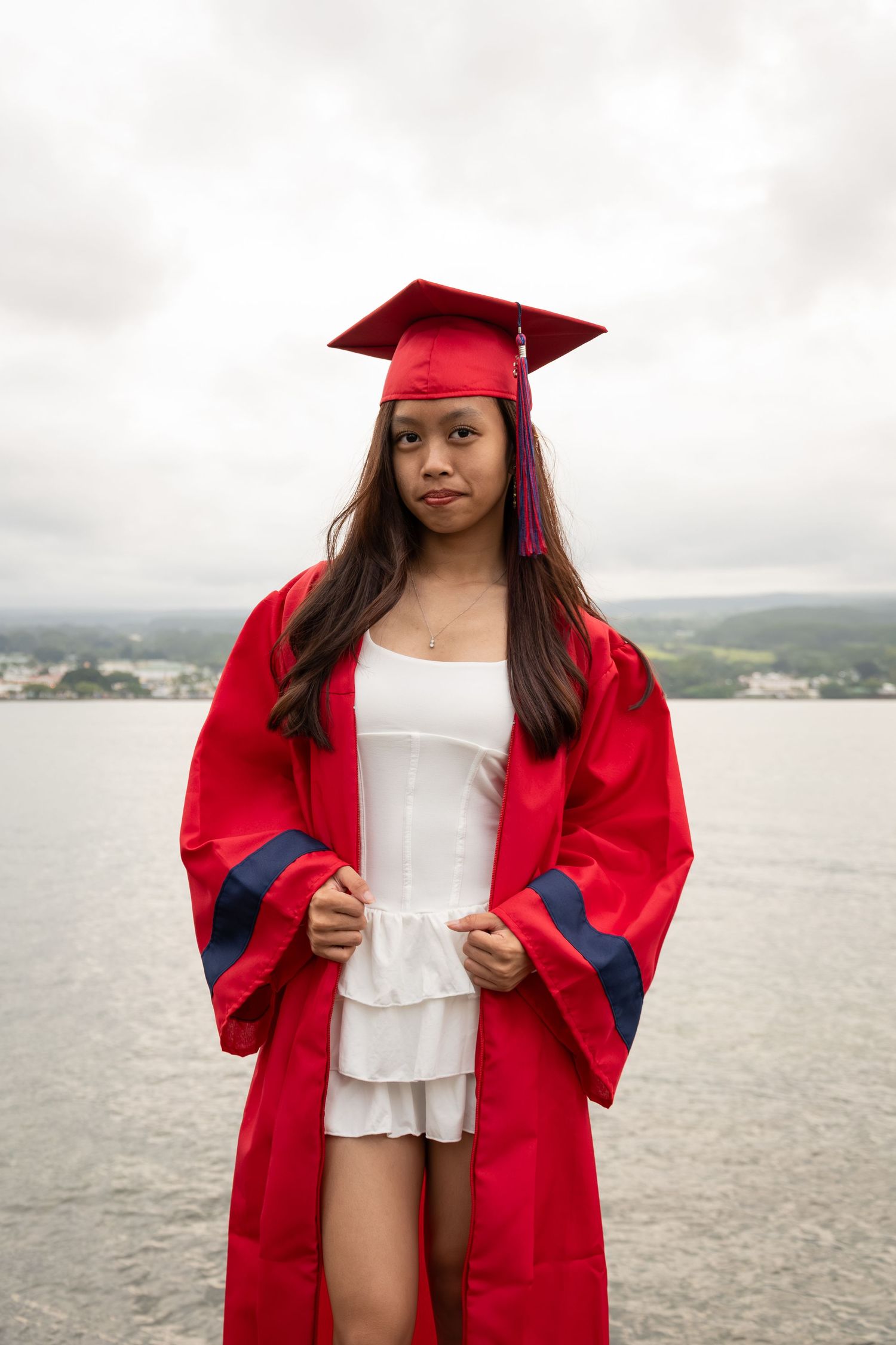 Young woman in red graduation cap and gown posing outdoors near water on a cloudy day.