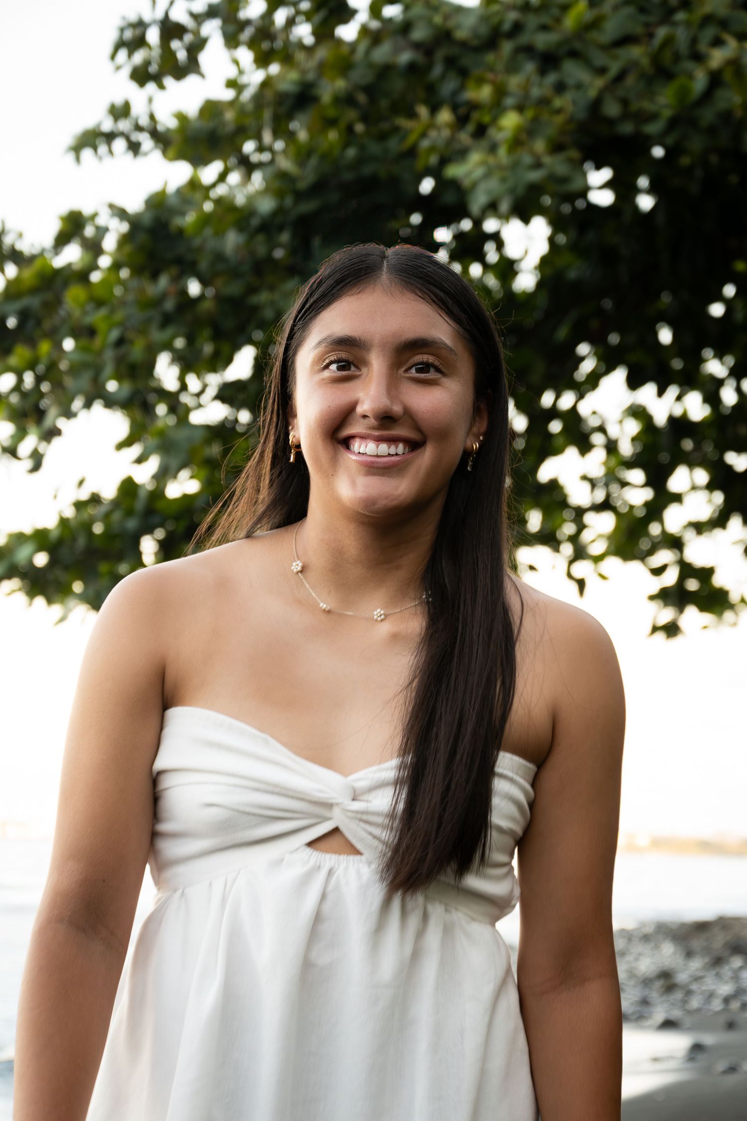 A person in a white sundress smiles outdoors with trees and a beach in the background.