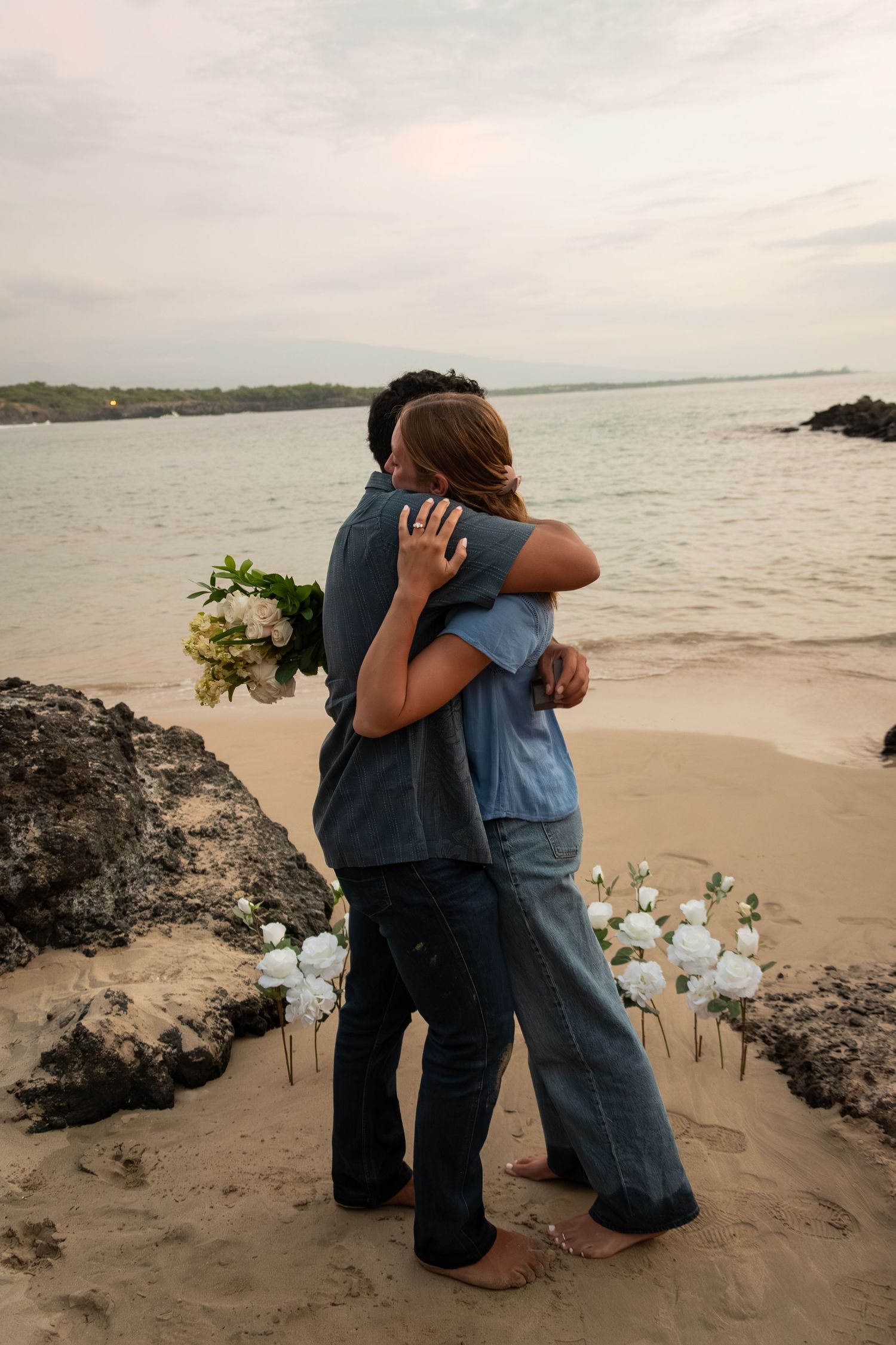 Couple embracing on a sandy beach at sunset, holding a white flower bouquet with floral decorations nearby.