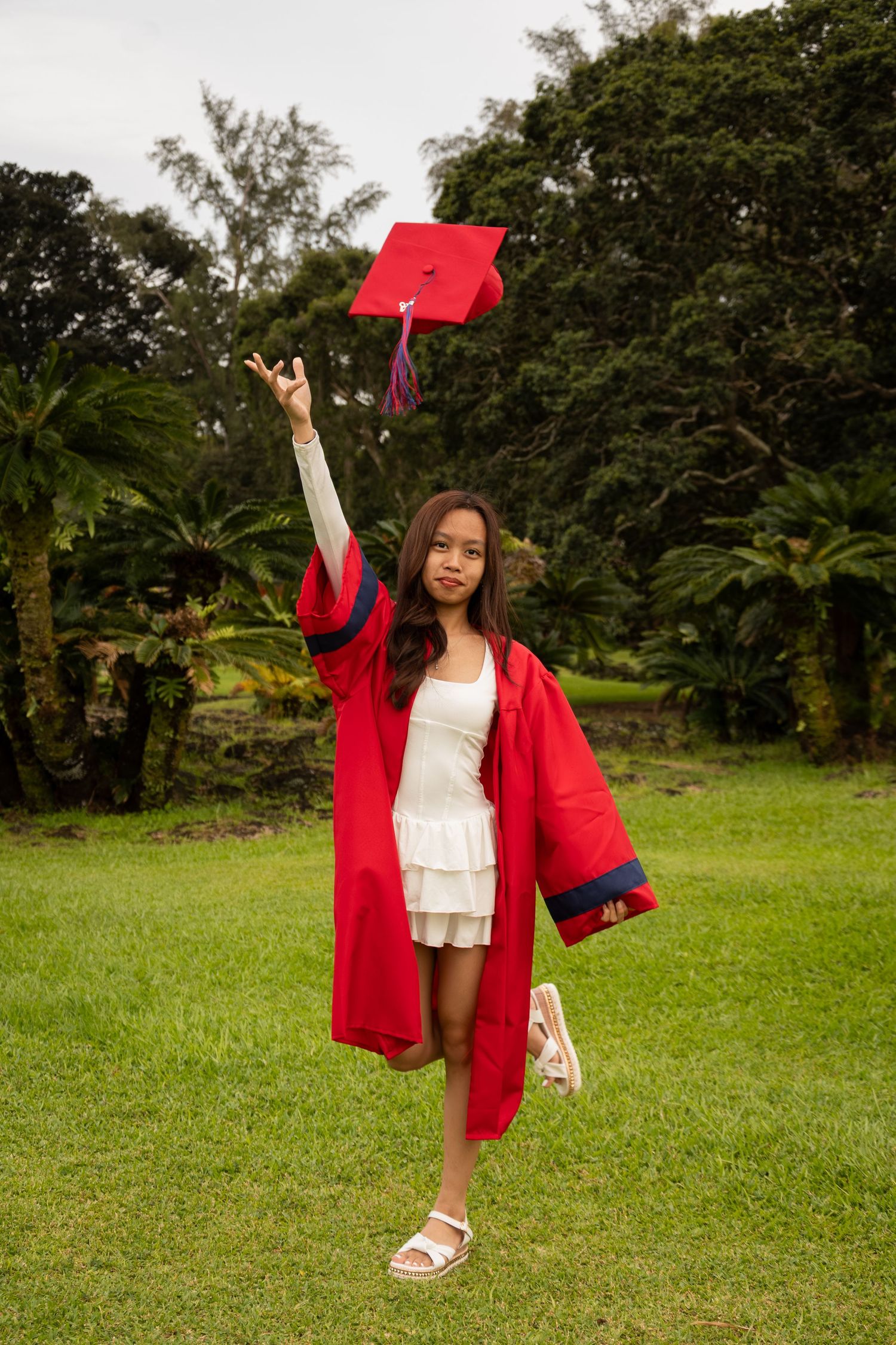 Young girl in red graduation cap and gown tossing her cap in the air outdoors on green grass.