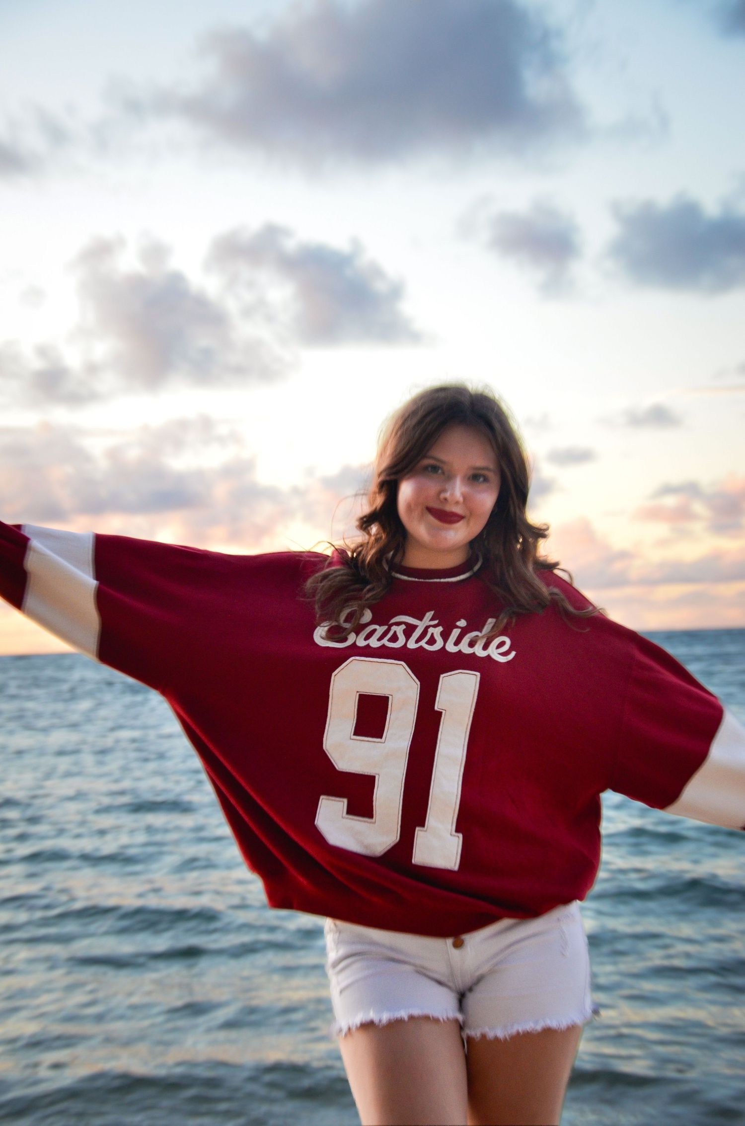 Person wearing red Eastside 91 jersey poses playfully at sunset by the ocean.