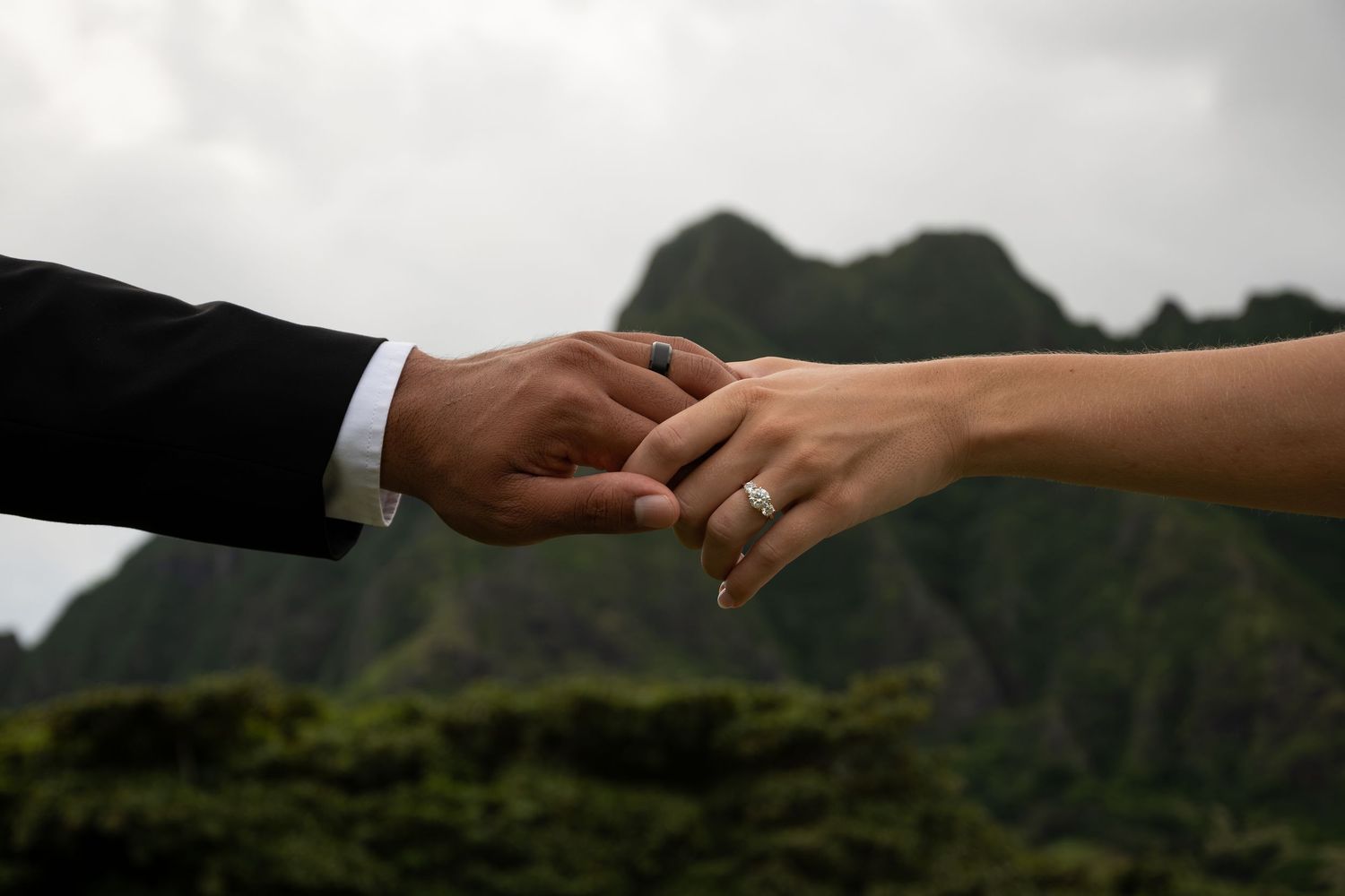 Newlywed couple holding hands with wedding ring visible, lush green mountains in background.