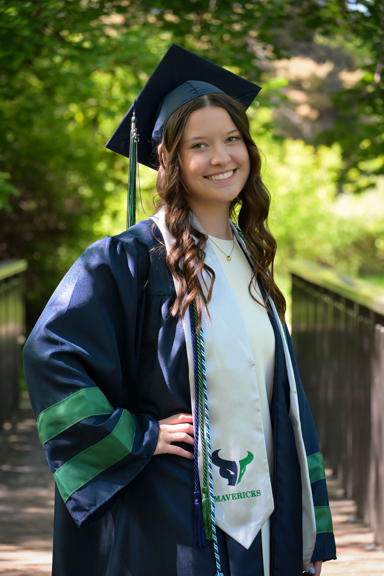 Graduate in navy cap and gown poses outdoors against greenery in a series of celebratory graduation photos.