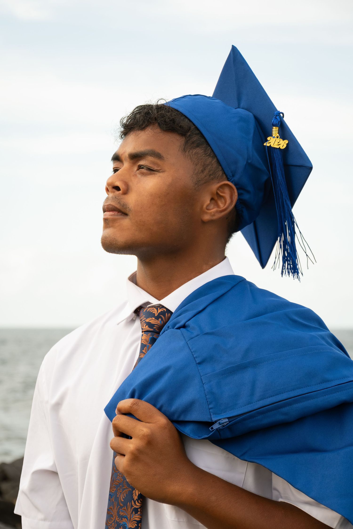 Young man in blue graduation cap and gown over white shirt and tie, gazing confidently with ocean backdrop.
