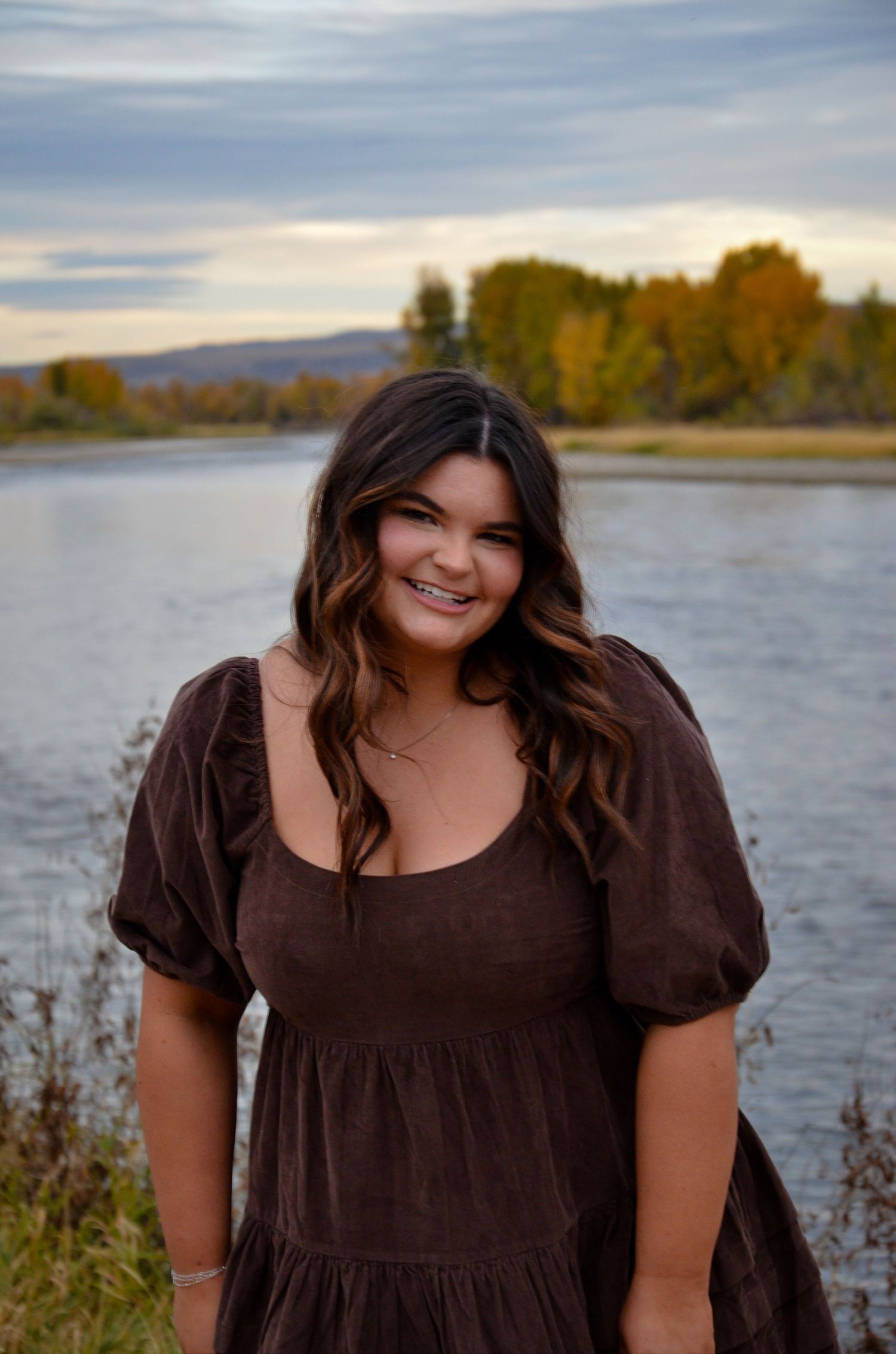 Person in brown dress poses by lake during autumn sunset.