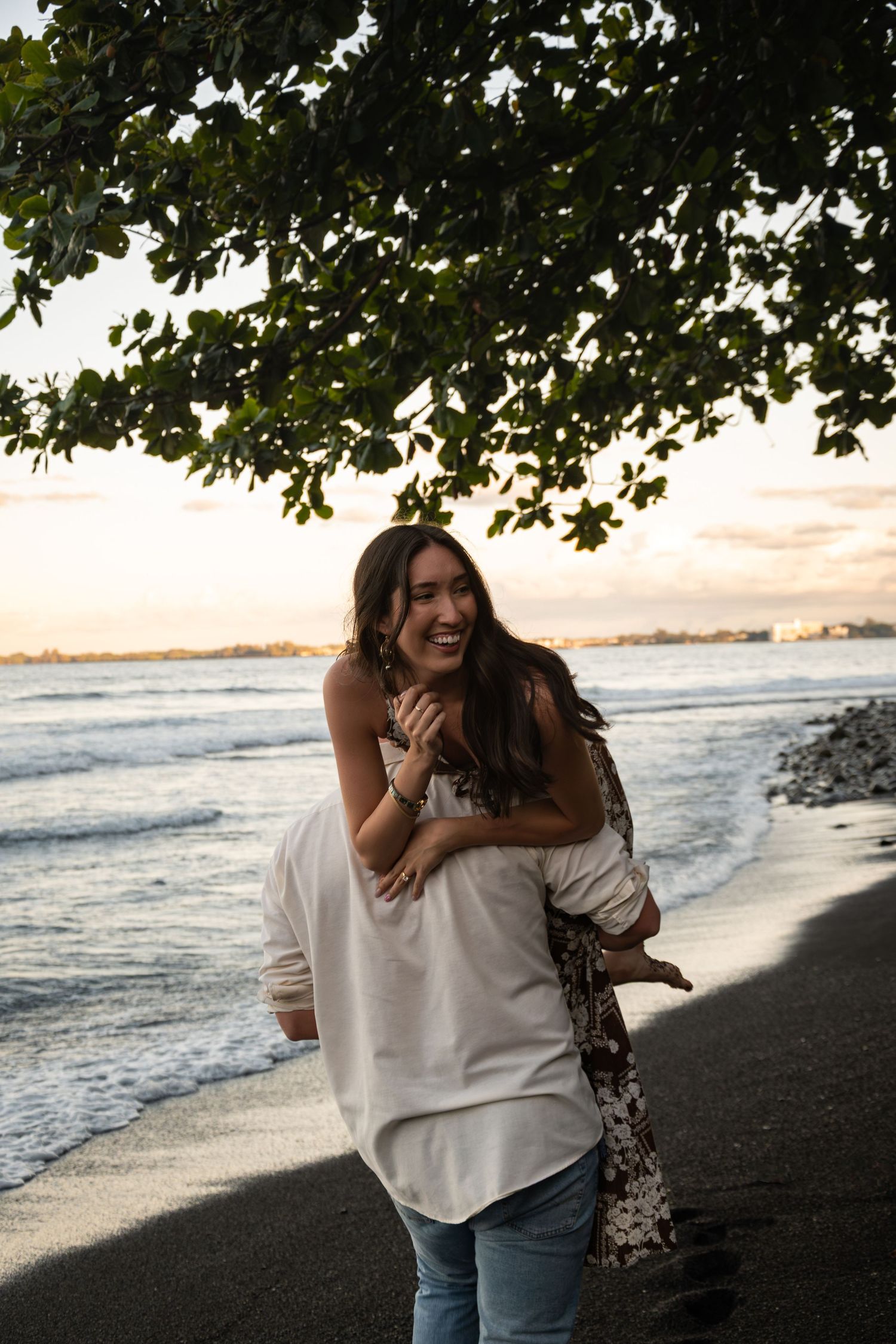 A joyful couple shares a playful piggyback moment on a black sand beach at sunset with ocean waves and trees overhead.