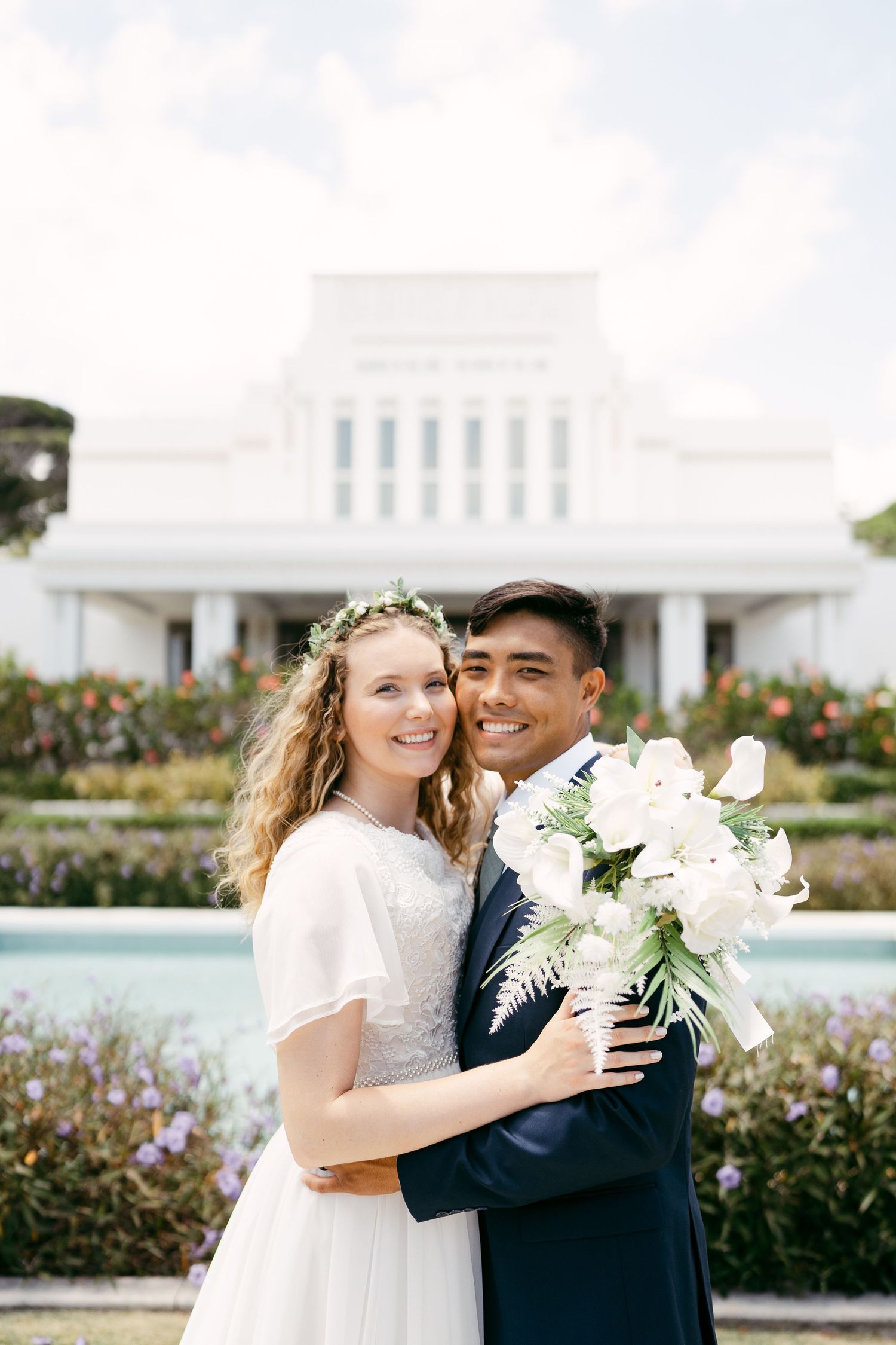A happy couple poses for their wedding portrait in front of a grand white temple with flowering gardens.