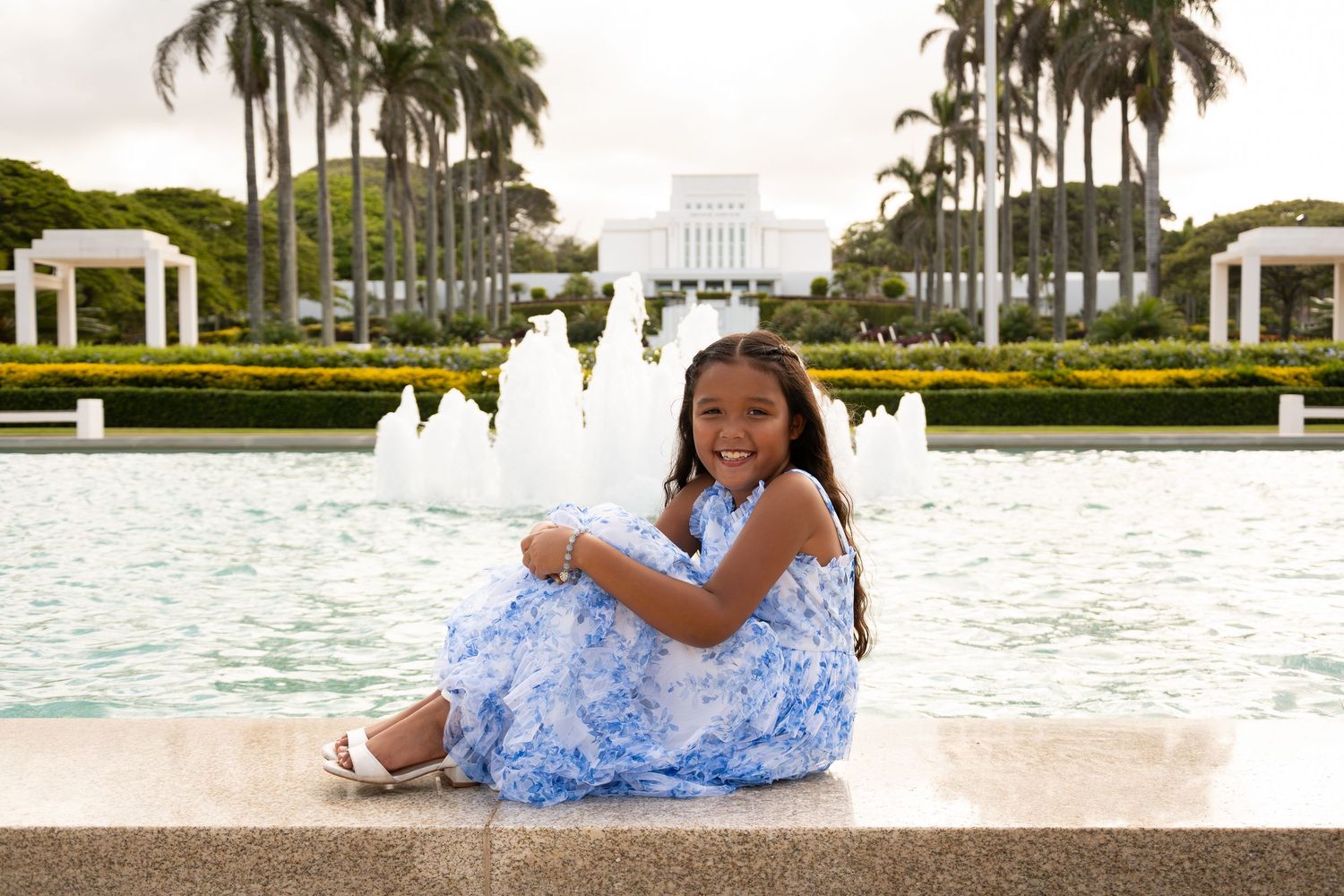 Young girl in blue floral dress sitting by fountain with palm trees and white temple building in background.