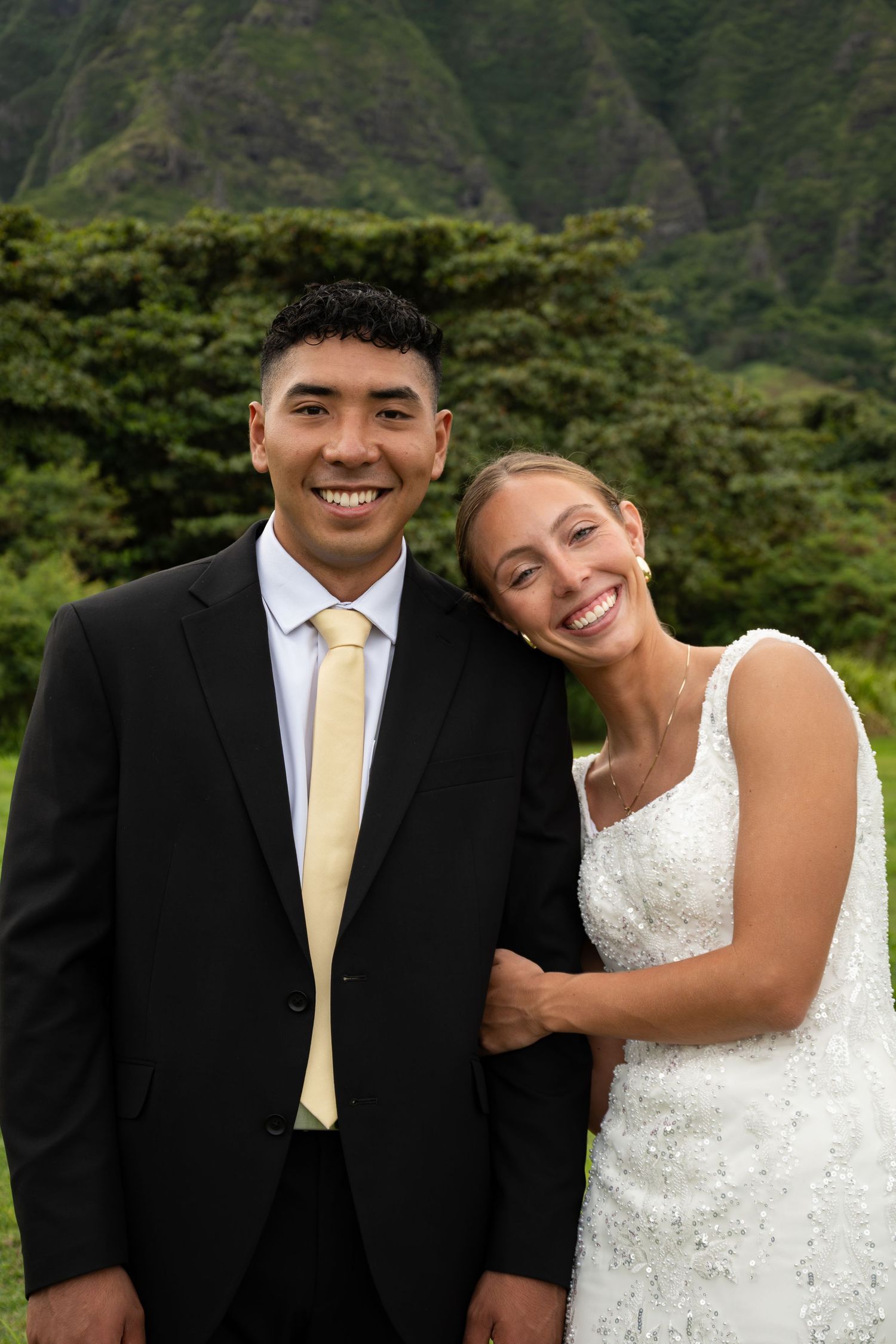 Smiling couple in wedding attire poses outdoors with lush green mountains in Hawaii background.