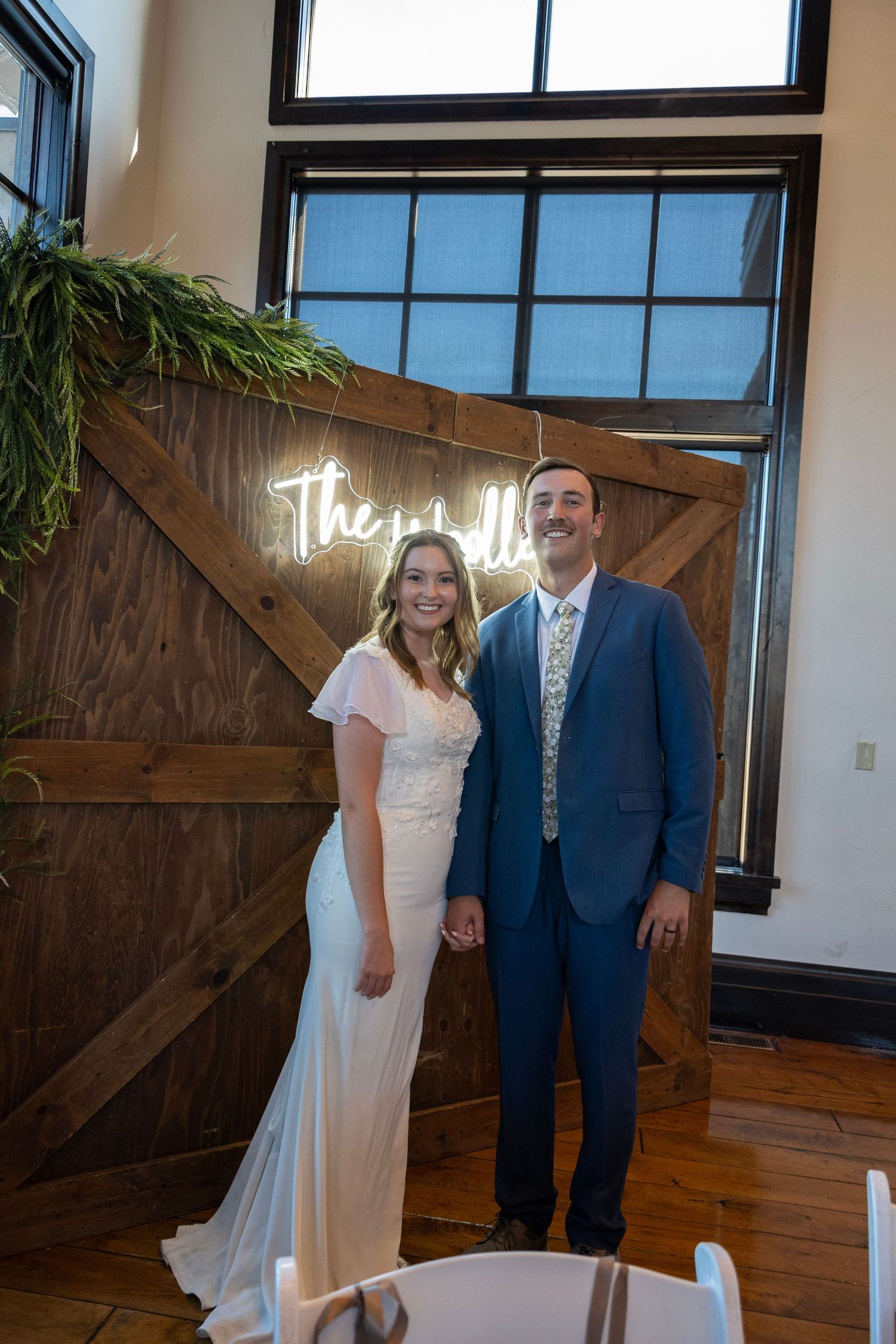 A wedding couple poses in formal attire in front of a rustic wooden barn door with neon signage.