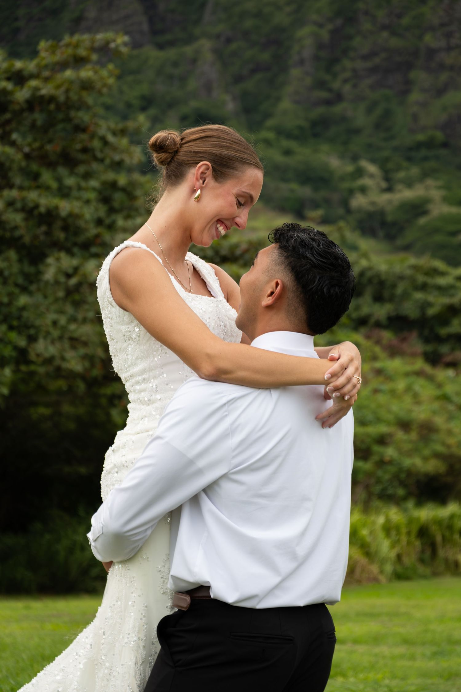 Groom lifting bride in white dress, sharing a joyful moment outdoors with lush green mountains behind them.