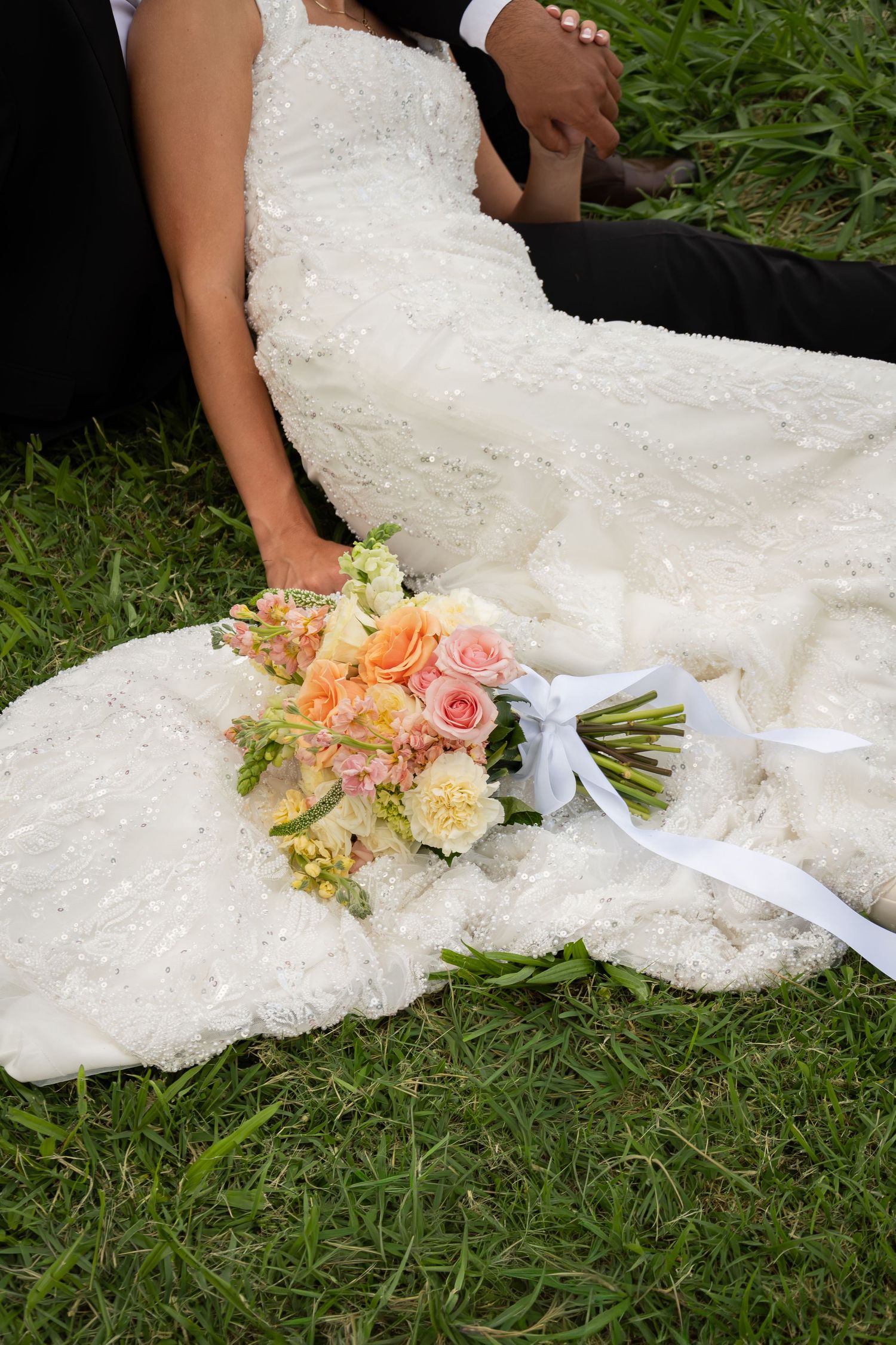 Bride and groom sitting on grass with beaded wedding dress spread out and floral bouquet with white ribbon beside them.