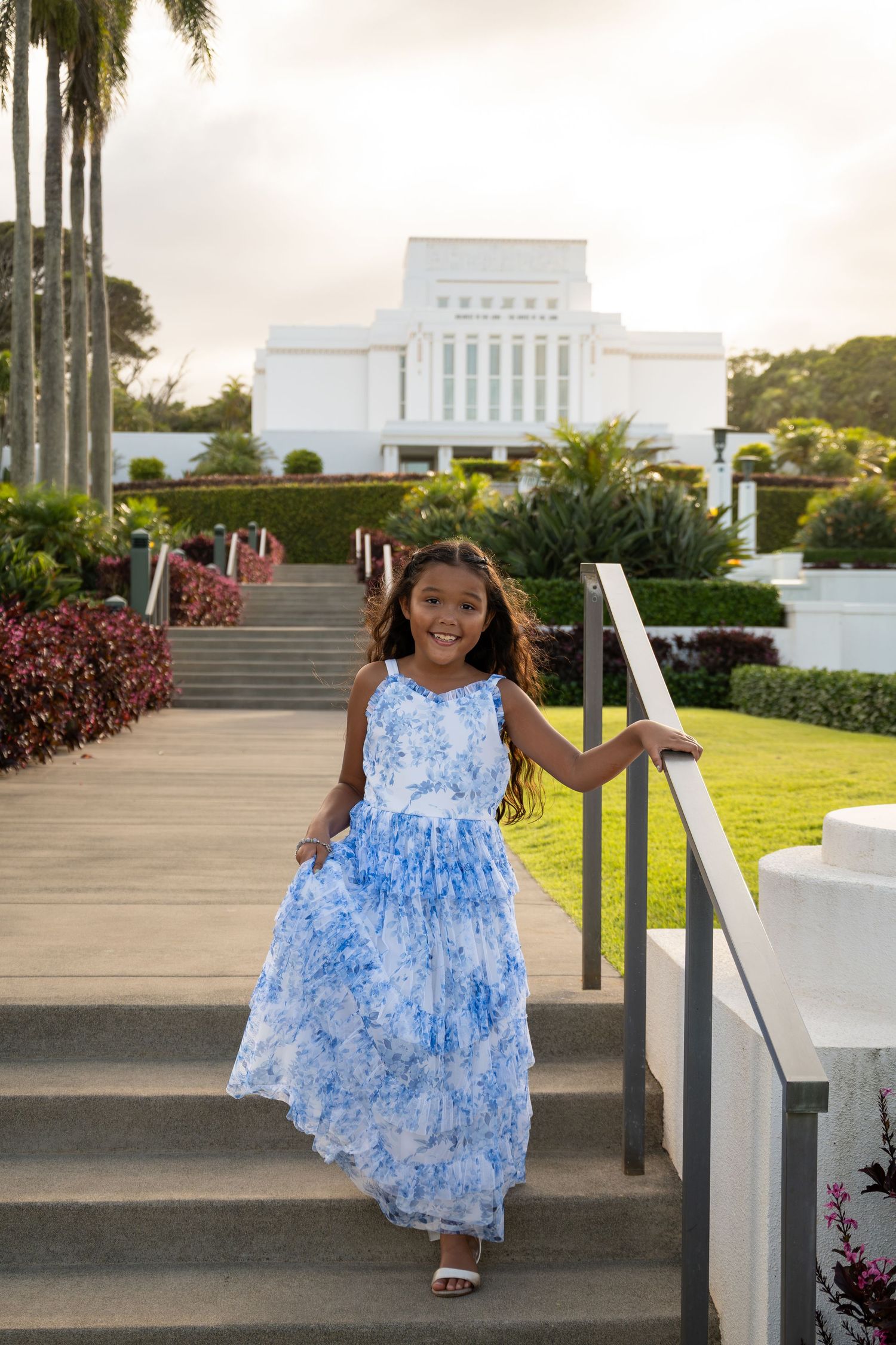 Young girl in blue floral dress walks up steps near a white temple building surrounded by lush tropical gardens.