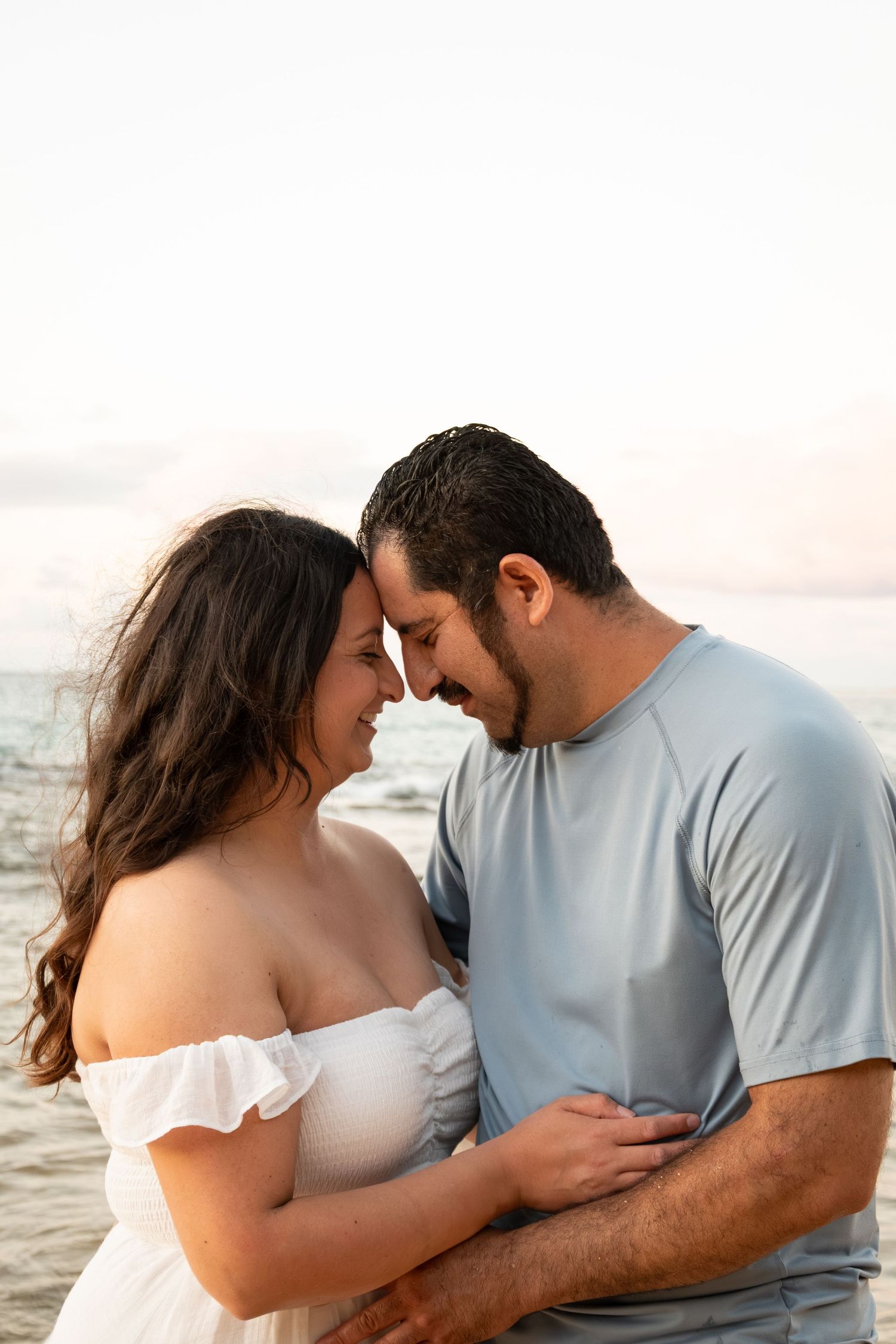 A romantic couple embraces on a beach at sunset, touching foreheads and smiling, wearing casual white and blue outfits.
