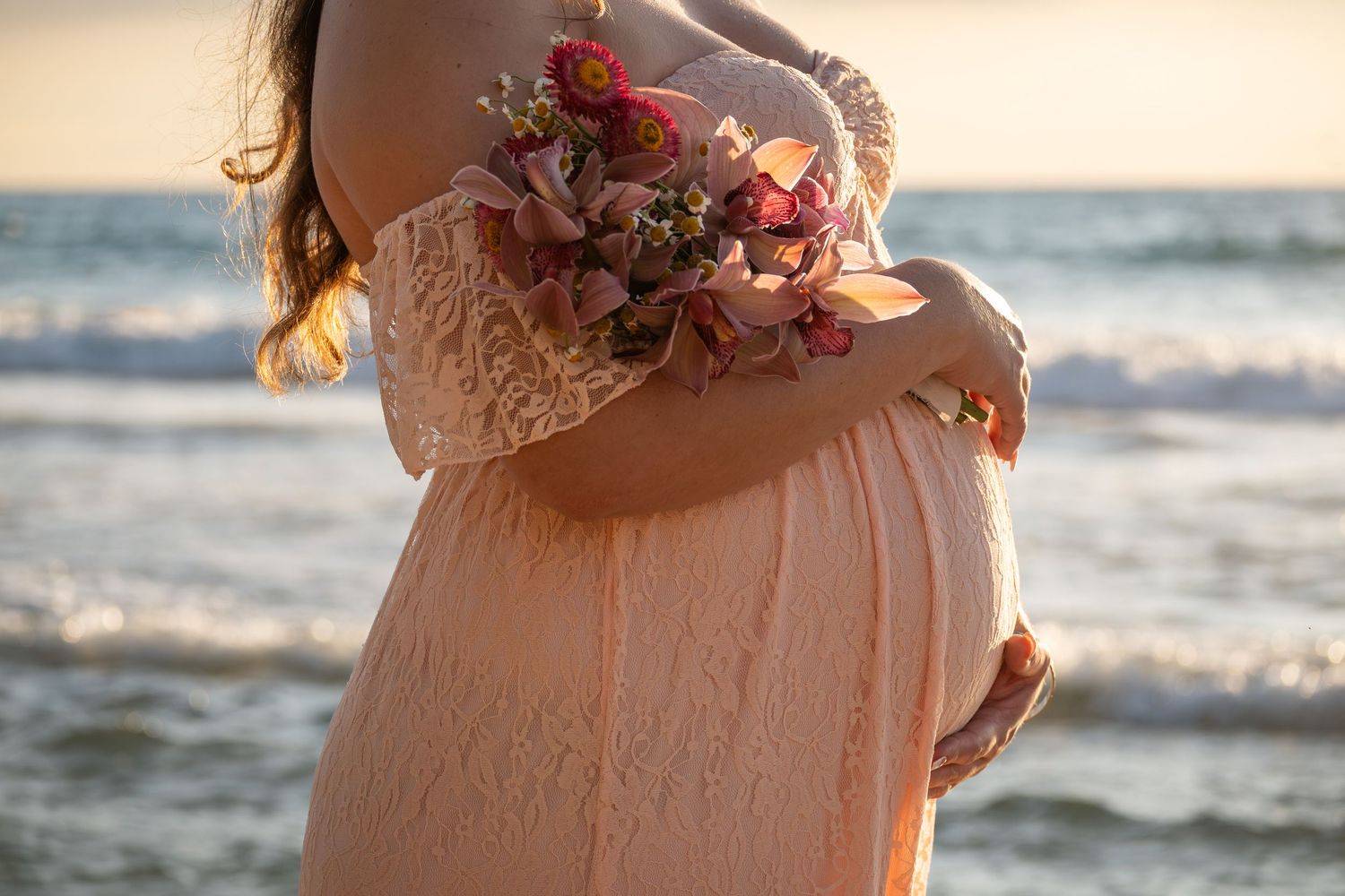 Pregnant woman in peach lace dress holding pink flowers at beach sunset, cradling baby bump by ocean waves.