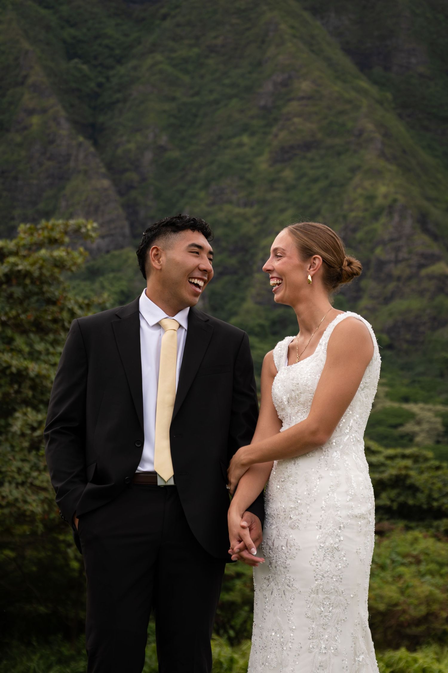 Smiling bride and groom hold hands laughing together with lush green Hawaiian mountains in the background.