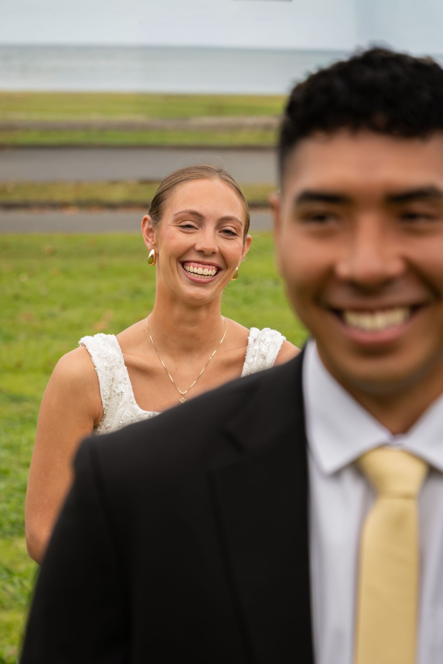 Smiling bride in white lace dress stands behind groom in black suit with gold tie at outdoor wedding.