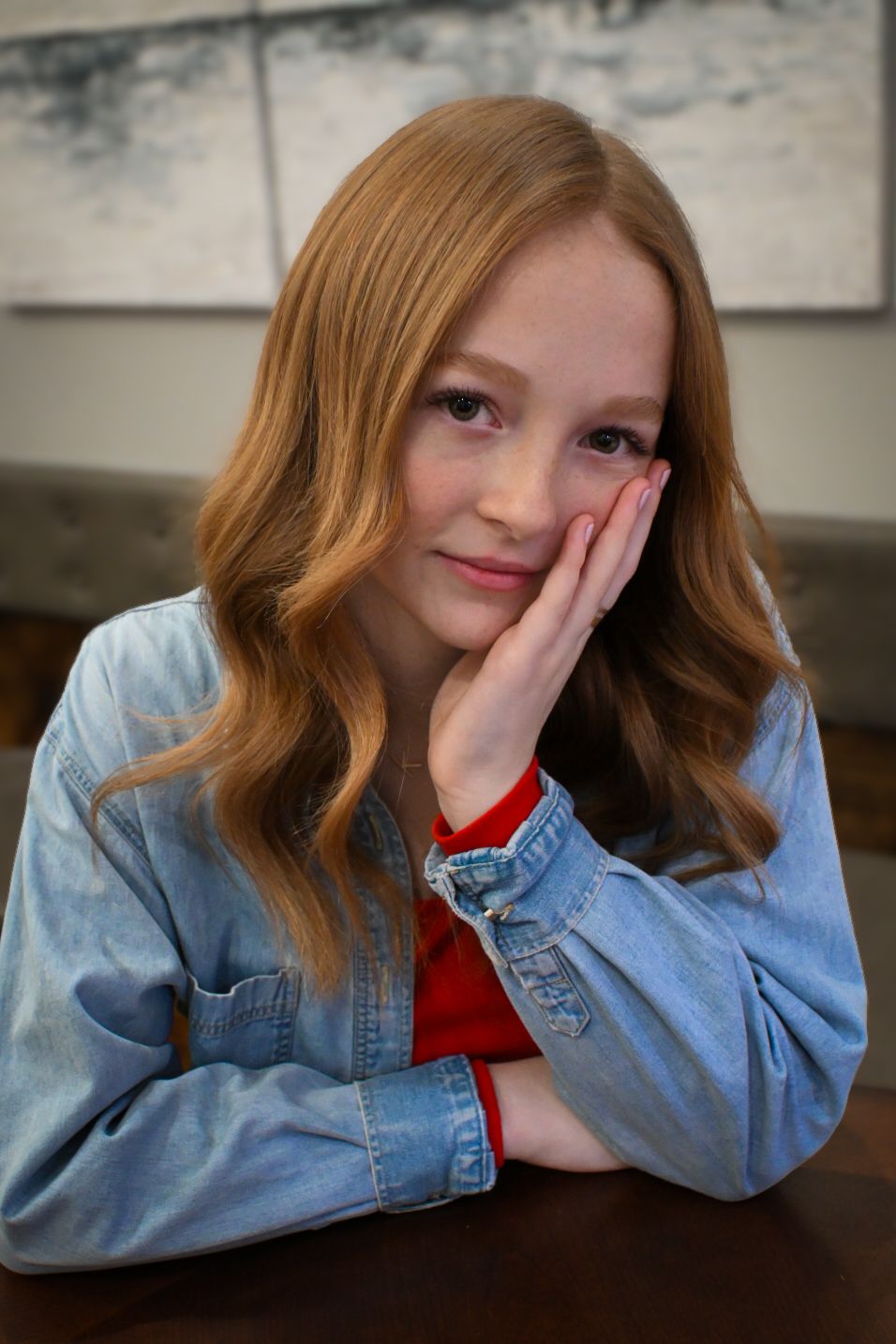 A person wearing a denim jacket and red top poses with hand against cheek in a casual indoor setting.