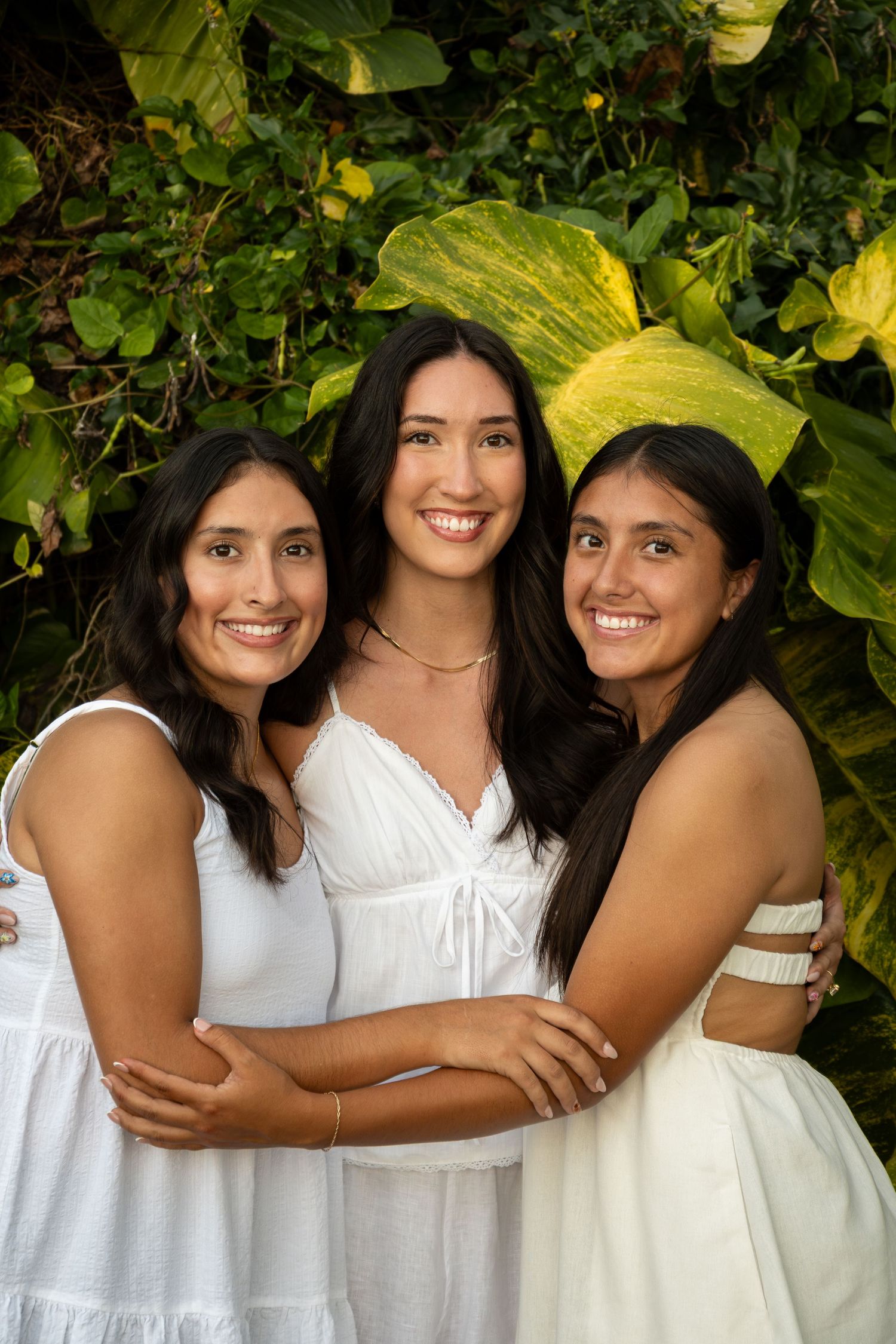 Three friends wearing white dresses embrace and smile together in a garden setting with lush green foliage.