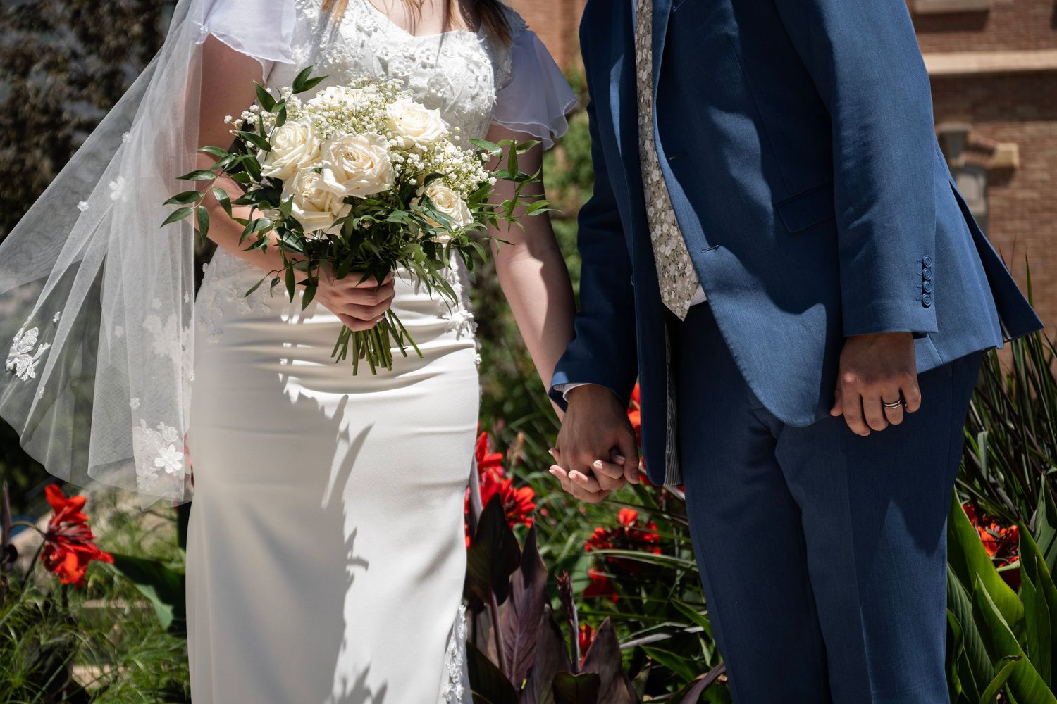 A wedding couple stands together outdoors, with the bride holding a white floral bouquet against her elegant white dress.