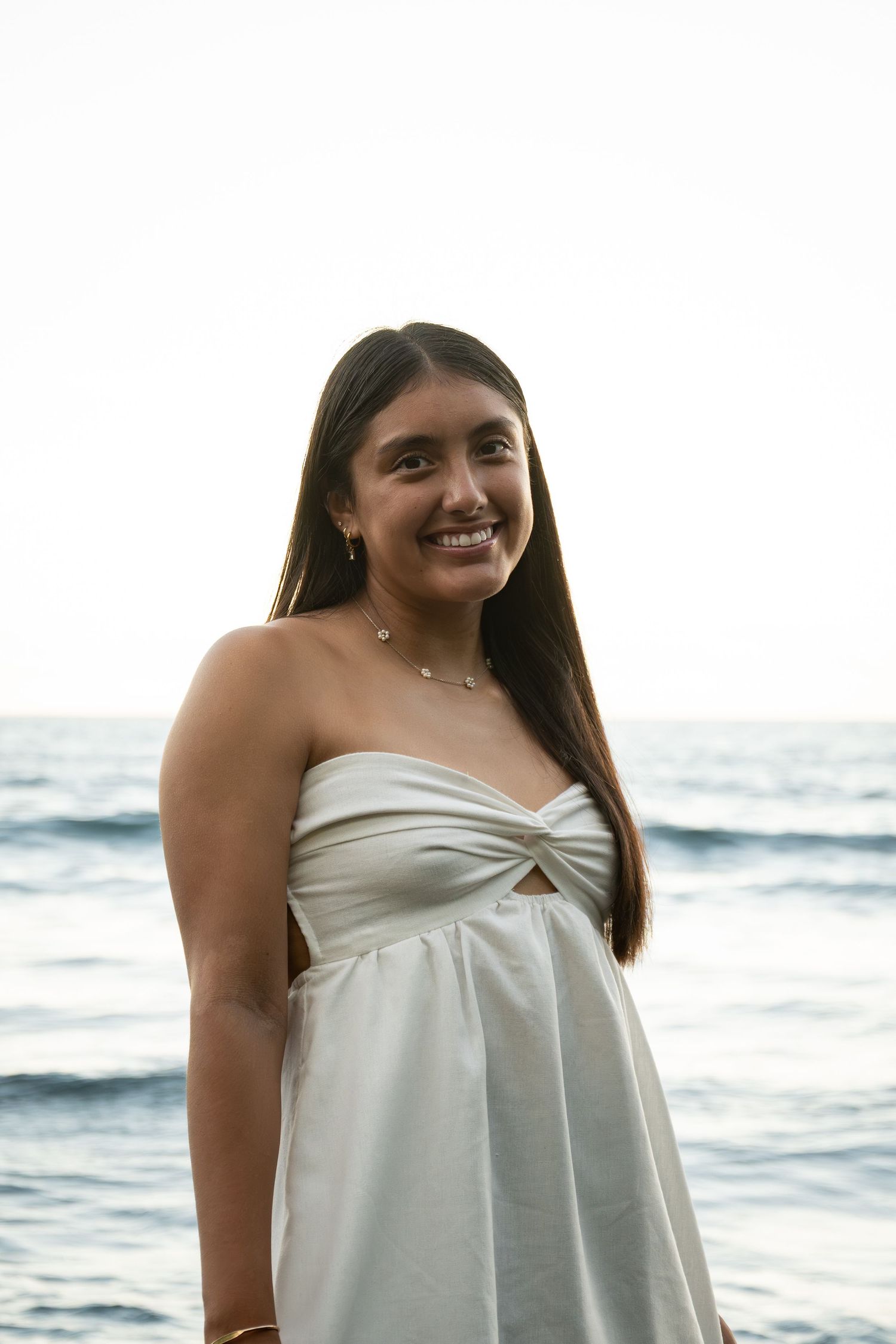 A person in a white strapless dress poses on a beach at sunset with ocean waves in the background.